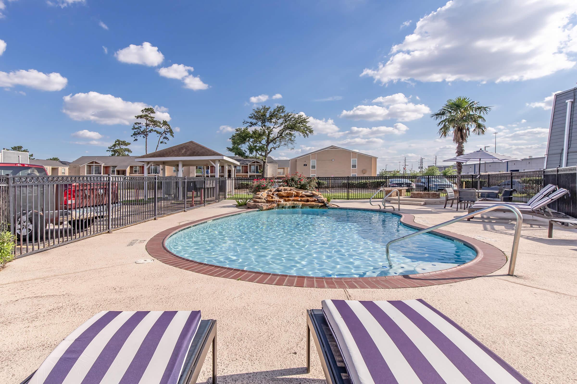 A serene swimming pool surrounded by lounge chairs and a fenced area. In the background, there are trees and residential buildings under a blue sky with fluffy clouds. A rock feature enhances the pool area, creating a relaxing outdoor atmosphere.