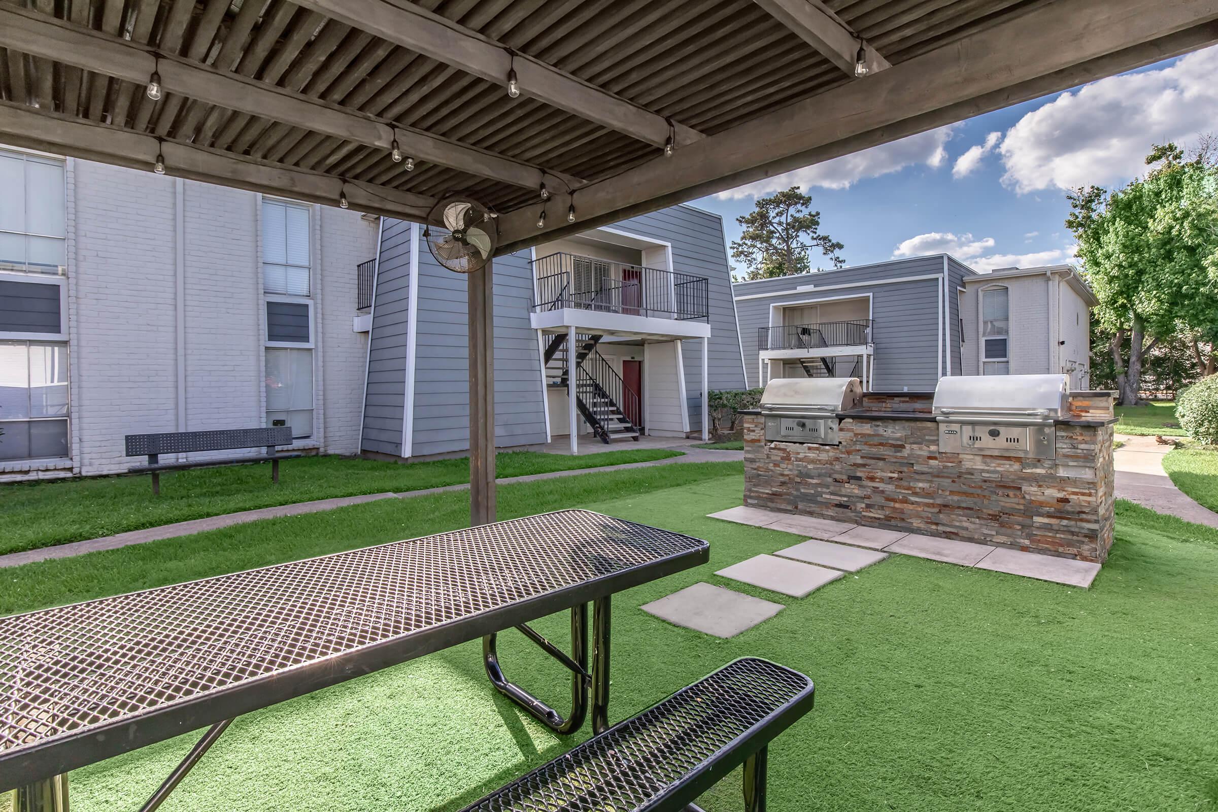 Outdoor seating area with a covered picnic table and a stone barbeque grill. The grassy space is surrounded by apartment buildings, featuring steps leading to upper-level units. The sky is partly cloudy, adding a scenic touch to the communal space.