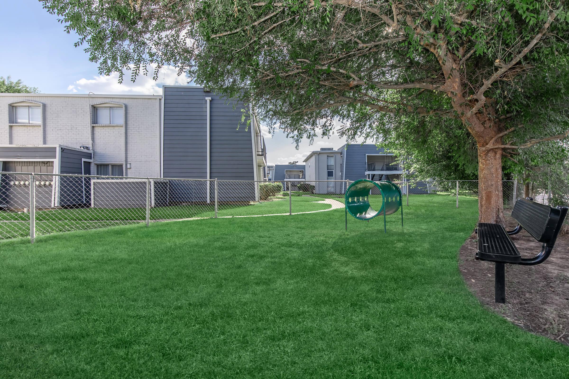 A grassy area in an apartment complex featuring a playground tunnel, benches, and trees. The background shows modern buildings with a fence enclosing the space, creating a serene environment for residents.