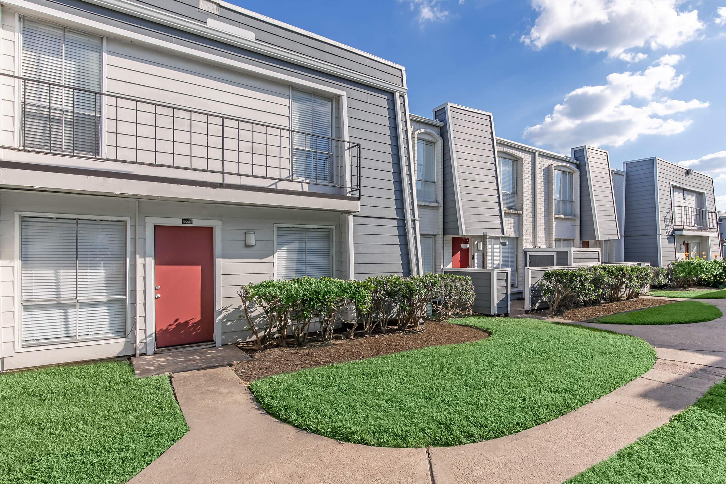 Exterior view of a modern residential complex with multiple two-story buildings. Each building has a light-colored exterior, large windows, and red doors. Lush green grass lines the pathways leading to the units, and there are well-maintained shrubs bordering the walkways under a partly cloudy sky.