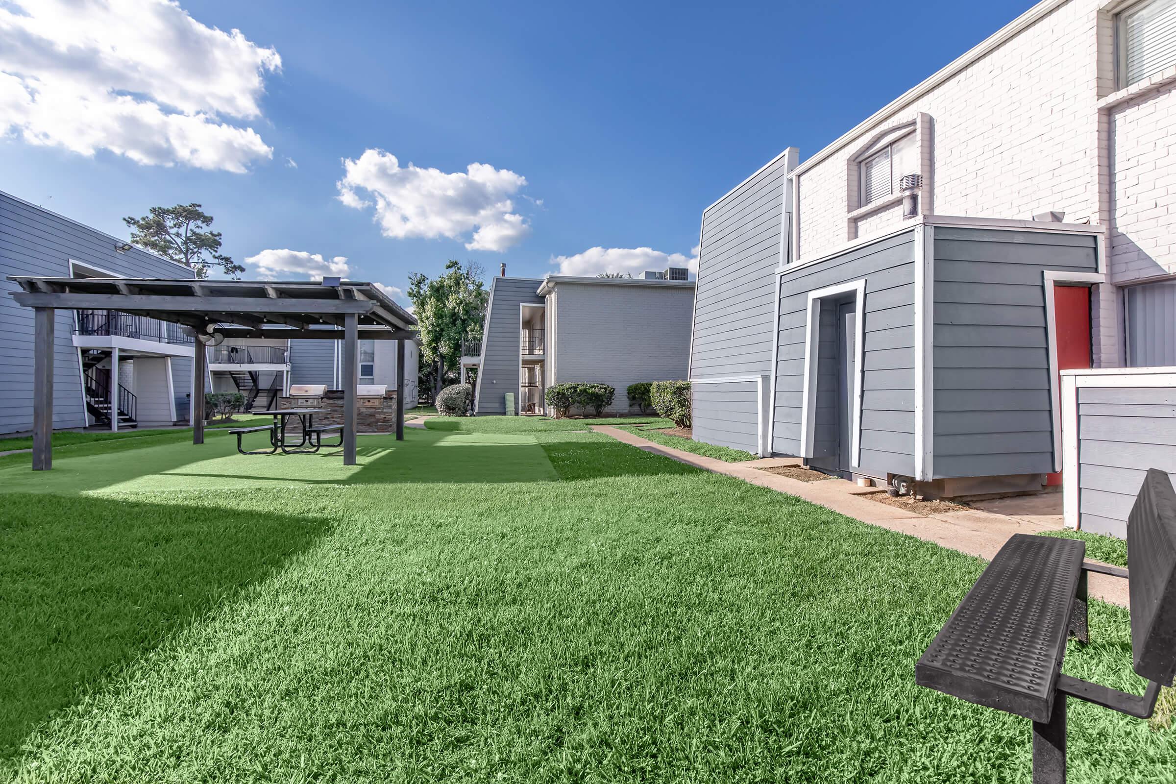 A well-maintained outdoor area featuring a lush green lawn, a shaded picnic gazebo, and nearby apartment buildings. The scene is brightened by a clear blue sky with a few clouds, creating a welcoming atmosphere for outdoor activities and relaxation.
