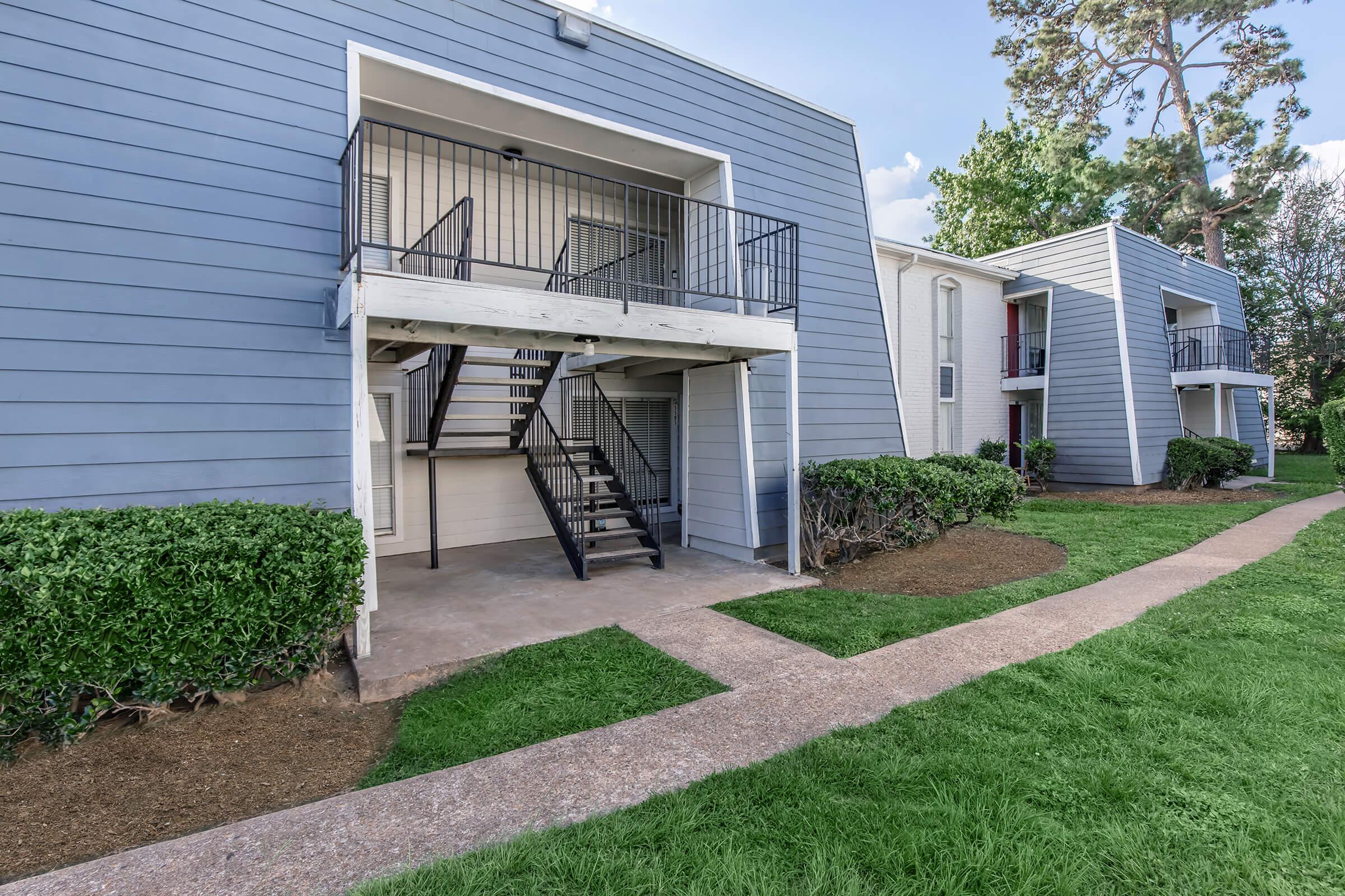 Exterior view of a modern apartment complex featuring two-tone blue and white siding. The building includes white railings and black metal staircases leading to upper-level units. Lush green grass and neatly trimmed bushes line the walkway, which leads to the entrance of the building.