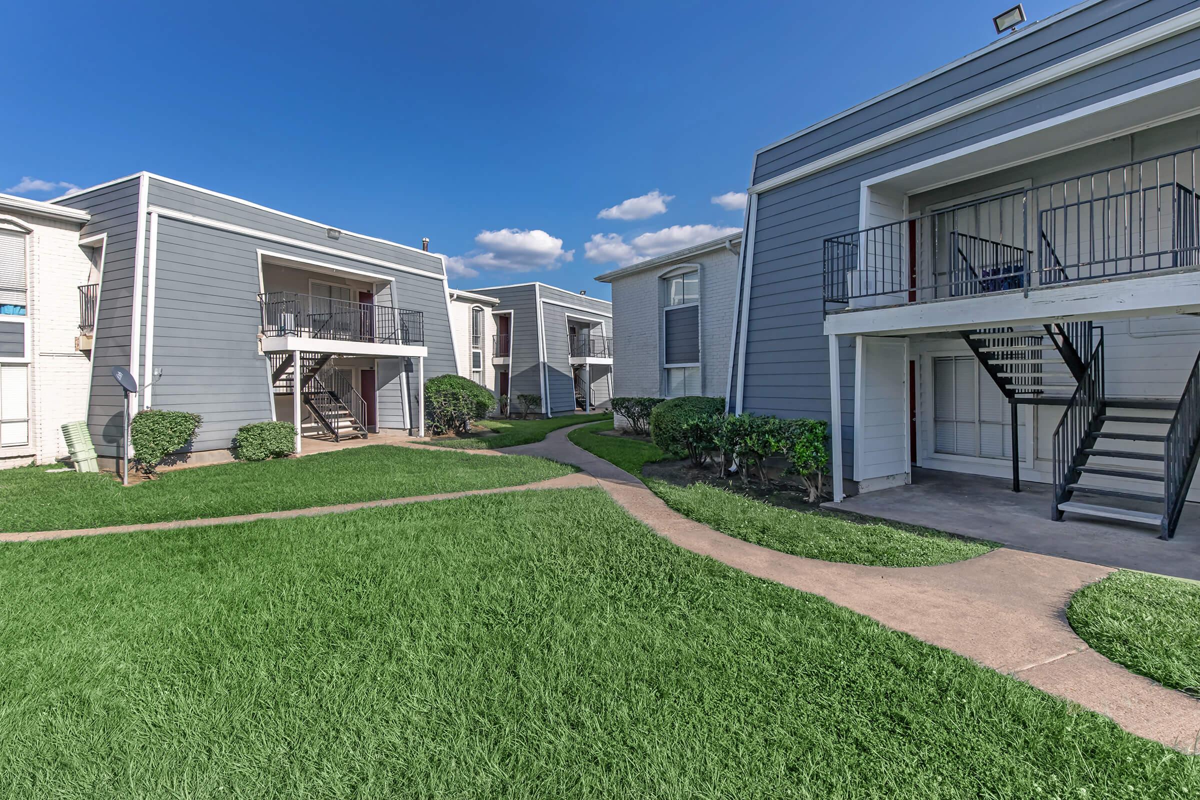 A view of an apartment complex featuring gray buildings with staircases, surrounded by well-maintained green grass and a curved pathway. The sky is blue with scattered clouds, creating a bright and inviting atmosphere.