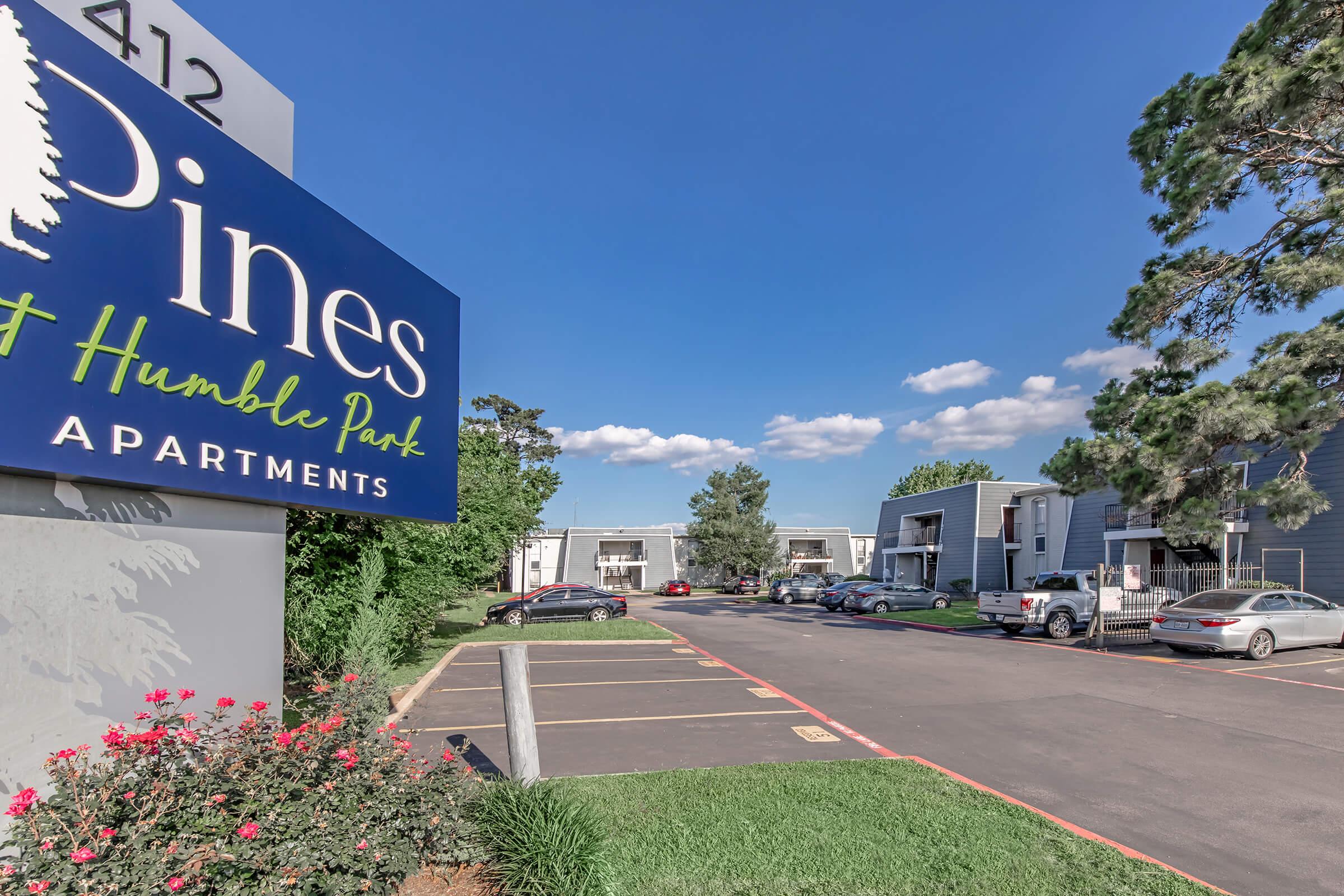 A sign for "The Pines at Humble Park Apartments" is prominently displayed, with a view of the apartment complex featuring trees and blue skies. The parking lot has several cars and well-maintained landscaping with flowers in the foreground.