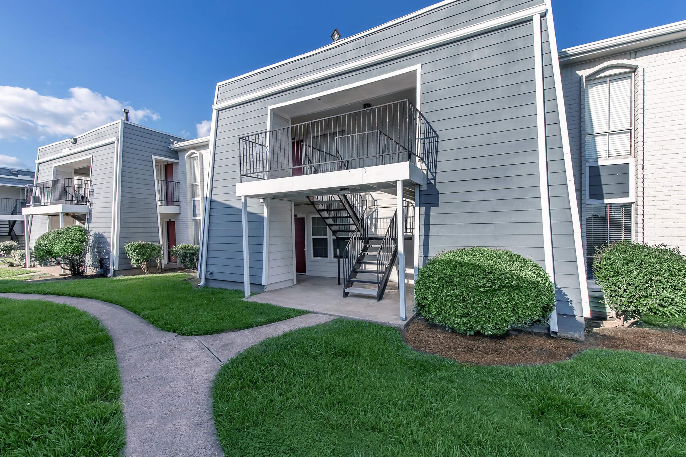 A modern two-story apartment building with a gray exterior. The building features a staircase leading to a balcony on the second floor. Well-manicured grass and shrubs surround the structure, and a paved path curves in the foreground, connecting the buildings. The sky is clear with a few clouds.