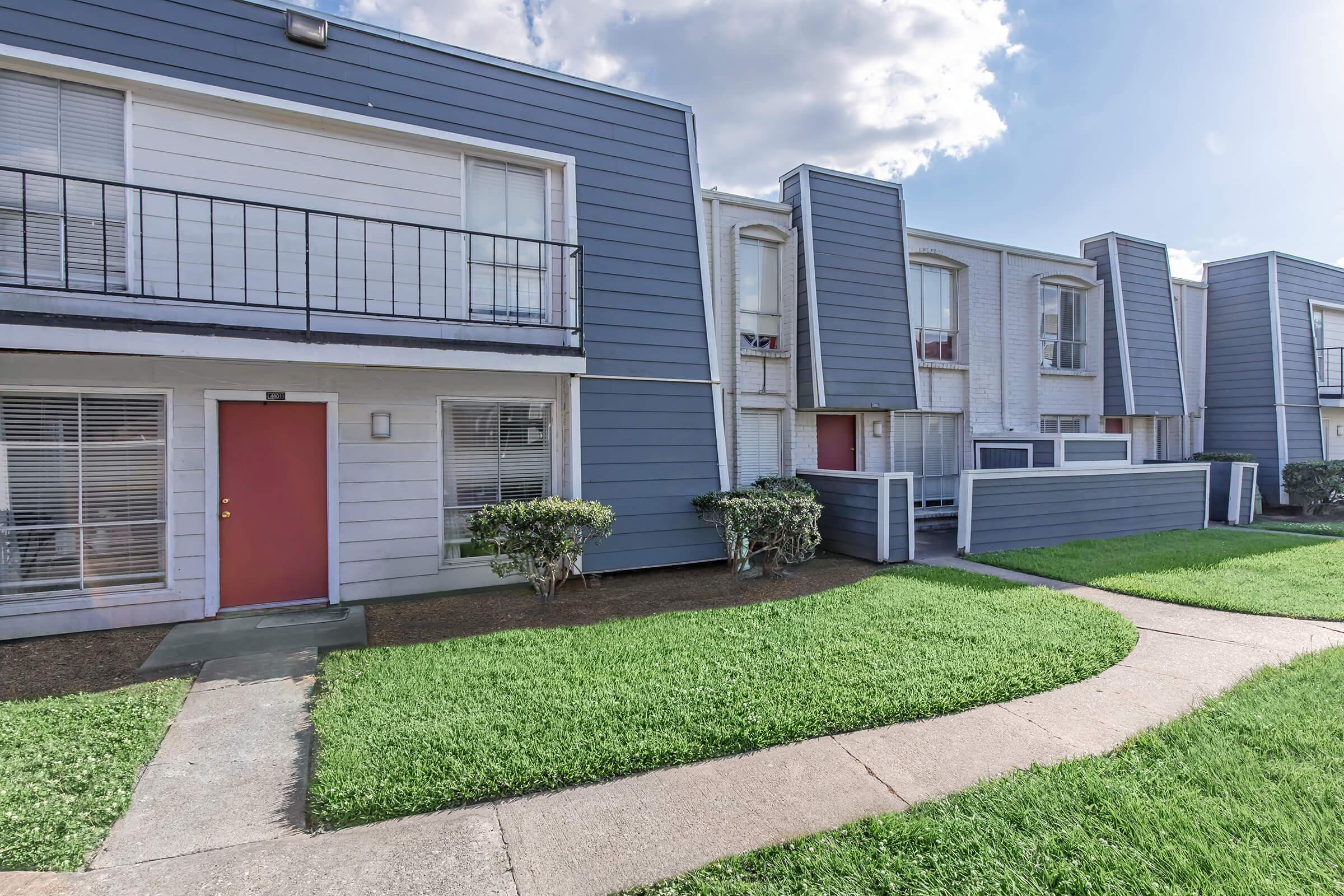 Exterior view of a multi-unit residential building featuring two stories, blue siding, and red doors. The landscape includes well-maintained green grass and a walkway leading to the entrance. There are balconies on the upper level, and the overall architecture has a modern design.