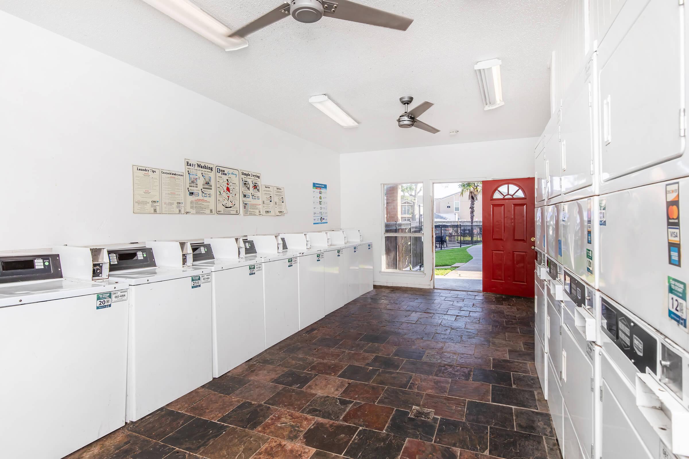 A clean laundry room with multiple white washing machines lined up against one wall. A bright red door leads to an outdoor area. The room has a ceiling fan and is well-lit with overhead lights. Informational posters are displayed on the wall, and the floor features dark, textured tiles.