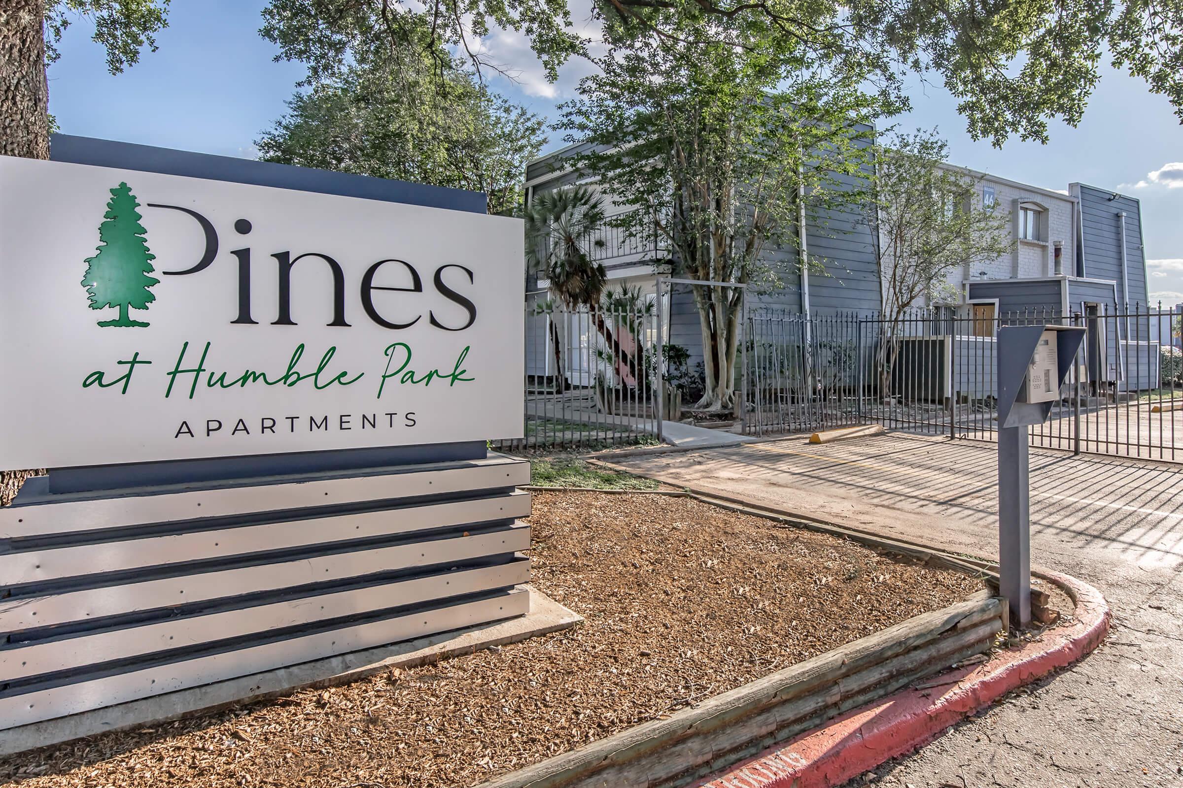 Sign for "Pines at Humble Park Apartments" with landscaping and a gated entrance visible. The apartment complex is surrounded by trees, providing a green setting. The scene is bright and inviting, suggesting a pleasant living environment.