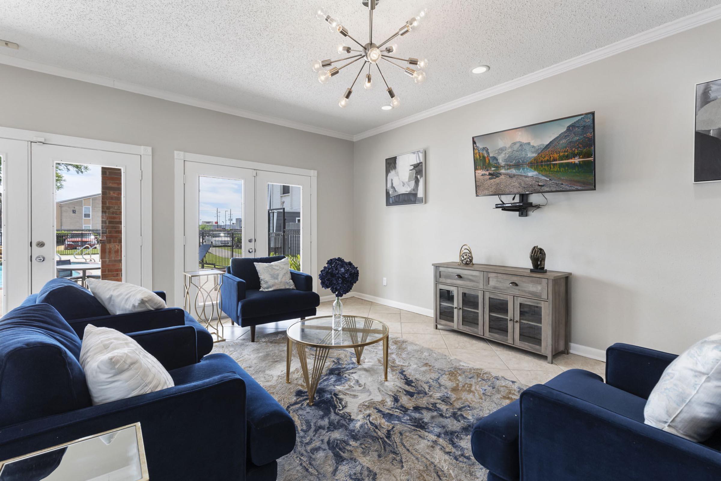 A stylish living room featuring a modern chandelier, a flat-screen TV mounted on the wall, a gray media console, and a round coffee table. Blue upholstered chairs and a sofa with decorative cushions offer comfort. Large windows allow natural light, enhancing the inviting atmosphere. Decorative artwork adorns the walls.
