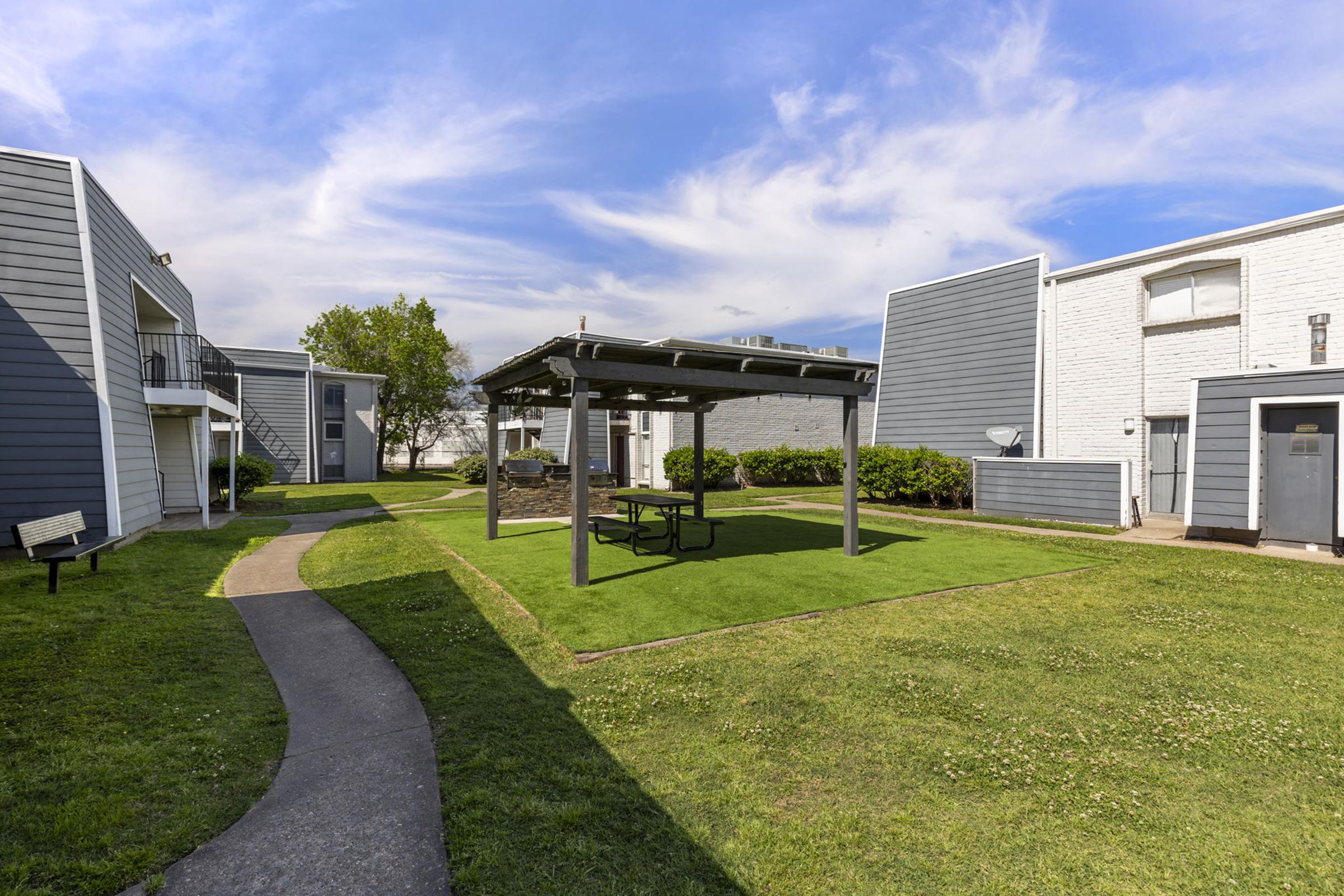 A grassy courtyard with a shaded picnic area under a wooden gazebo. Surrounding the courtyard are multi-story apartment buildings with gray siding. A paved walkway curves through the area, leading past a bench and greenery under a partly cloudy blue sky.