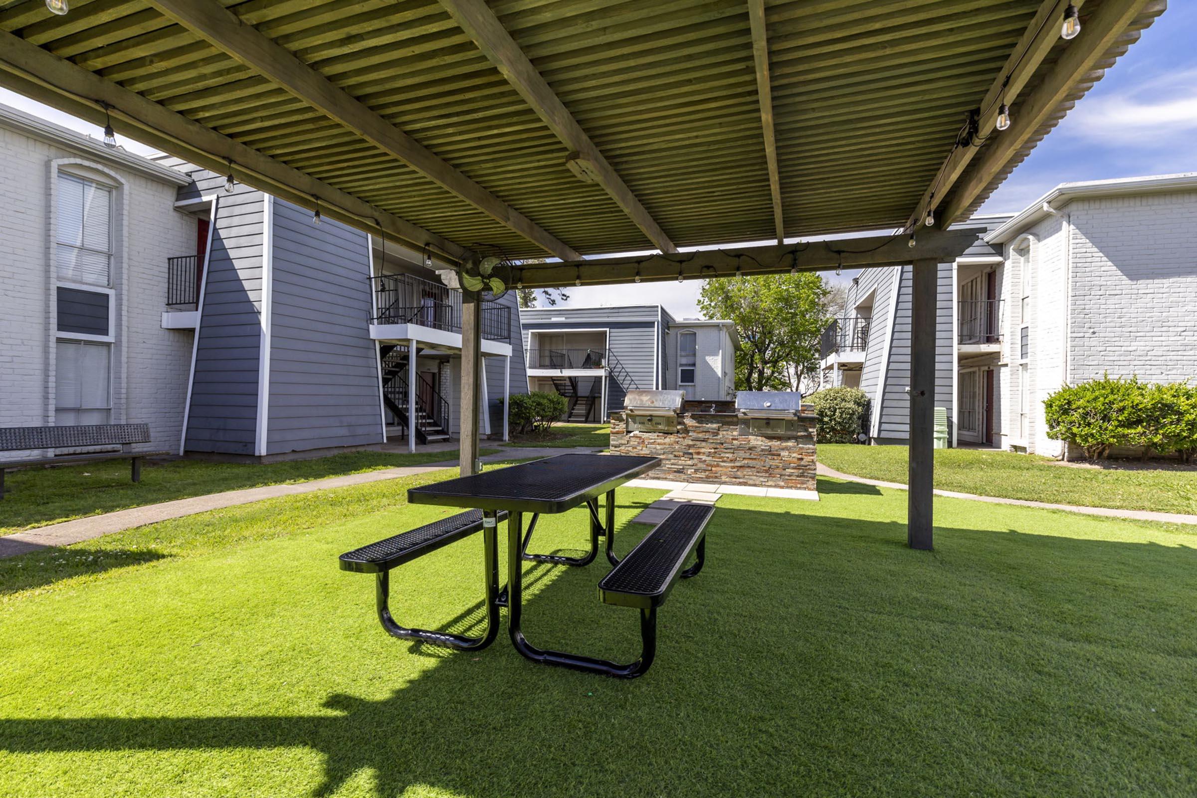 A shaded picnic area featuring a black metal table with attached benches, situated on green grass. Surrounding the area are buildings with gray and white exteriors. The sky is partly cloudy, and there are decorative trees in the background.
