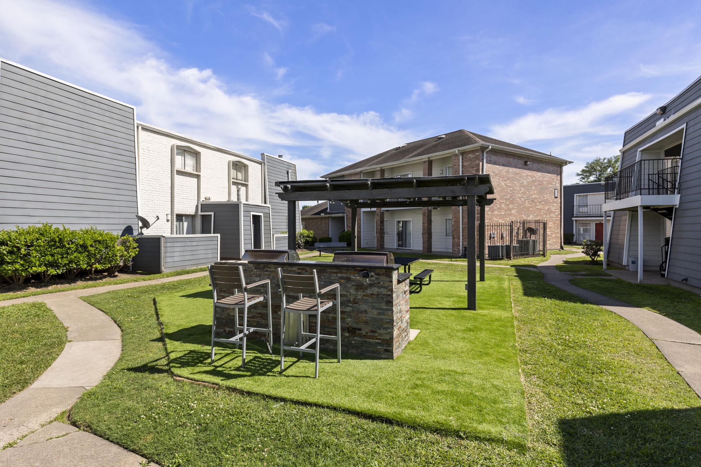 Outdoor courtyard featuring well-maintained green lawns, a barbecue area with stone structure and seating, surrounded by gray apartment buildings under a clear blue sky. Pathways meander through the space, inviting residents to enjoy the communal area.