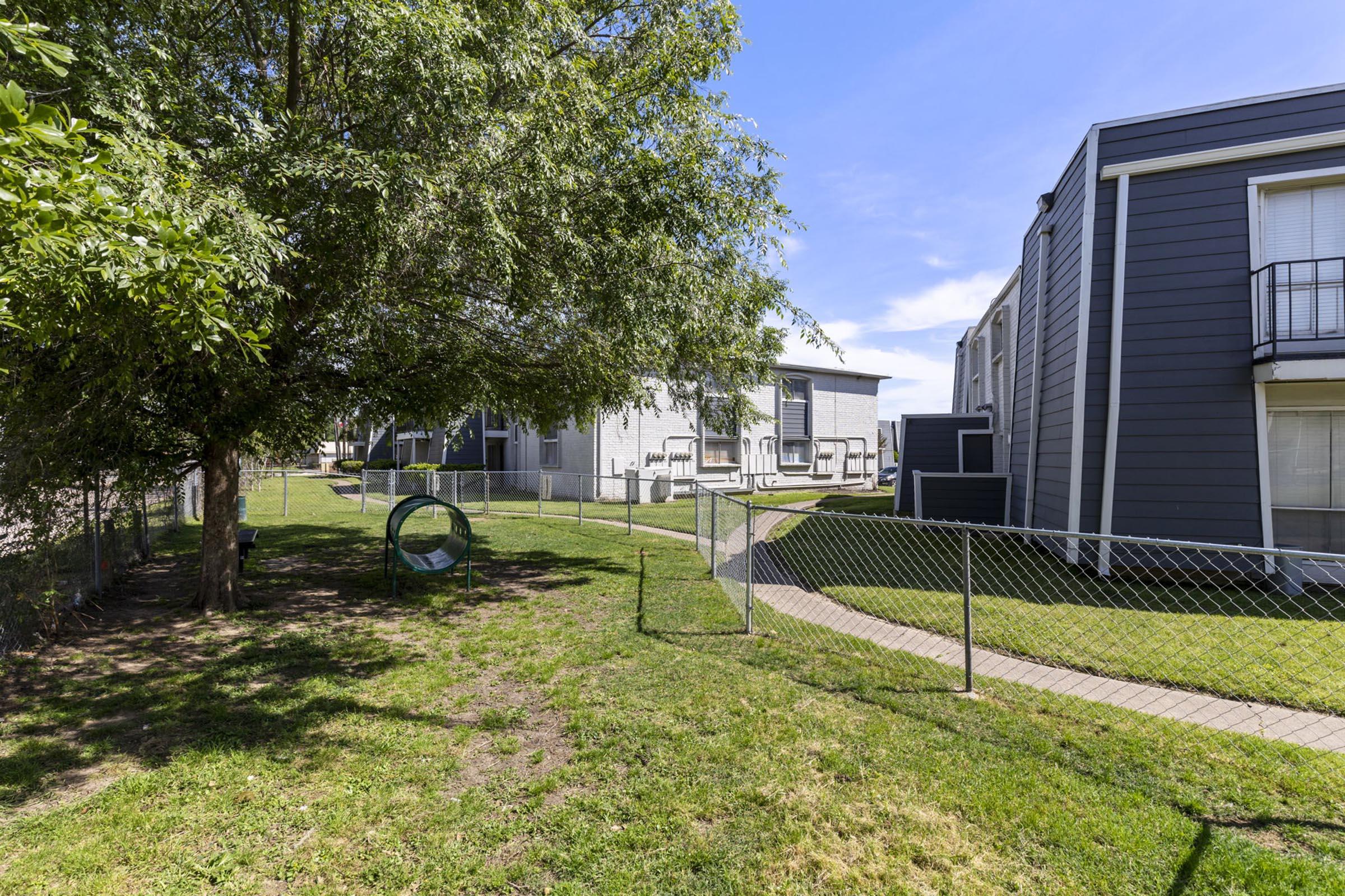A grassy area between buildings with a chain-link fence. There is a tree providing some shade, a circular play structure in the foreground, and pathways leading through the yard. The sky is clear with a few clouds, and the buildings are modern residential structures.