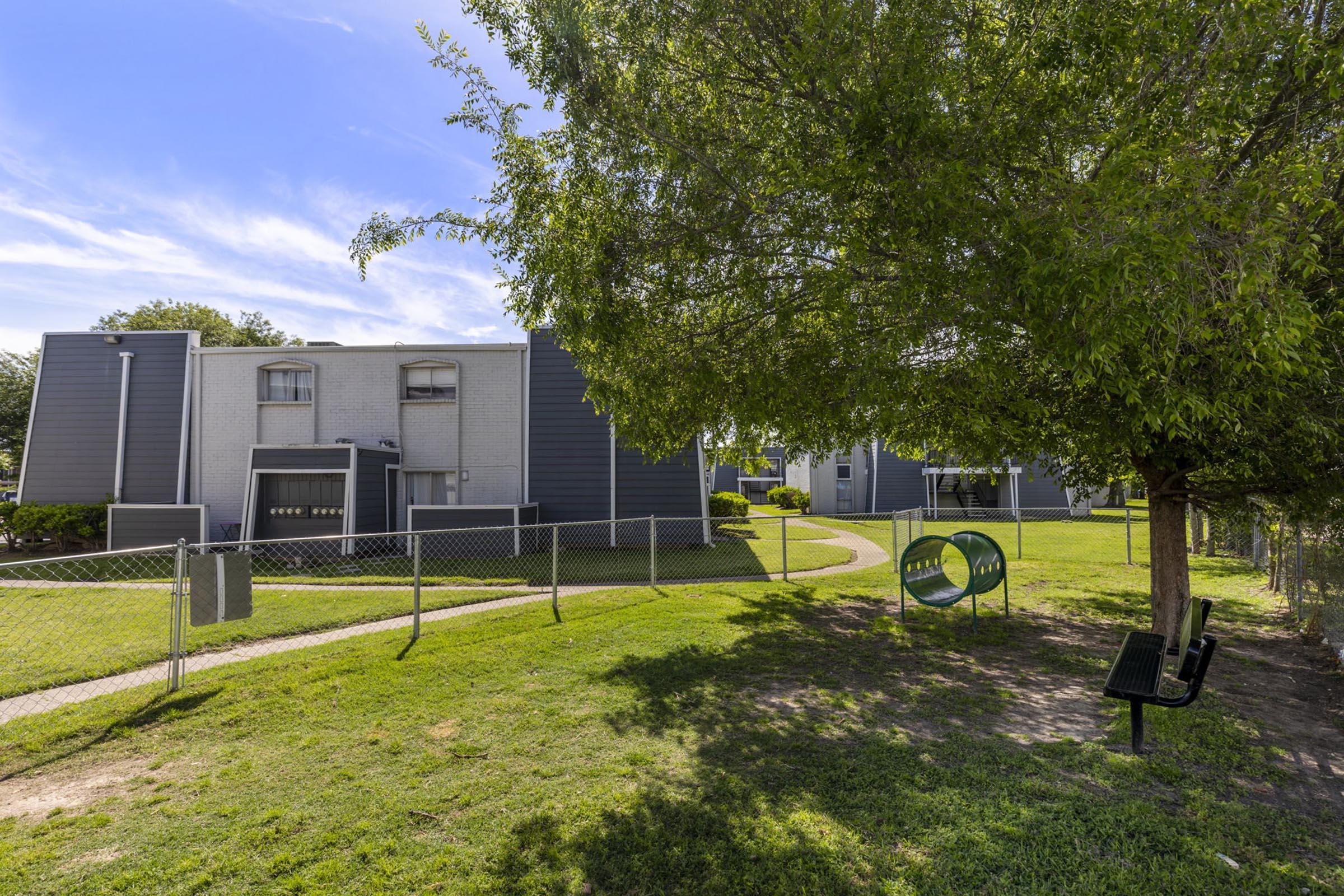 A peaceful residential area featuring two-story apartment buildings surrounded by green grass. A wooden bench under a tree is near a fence-lined pathway. Lush foliage and clear skies create a tranquil atmosphere.