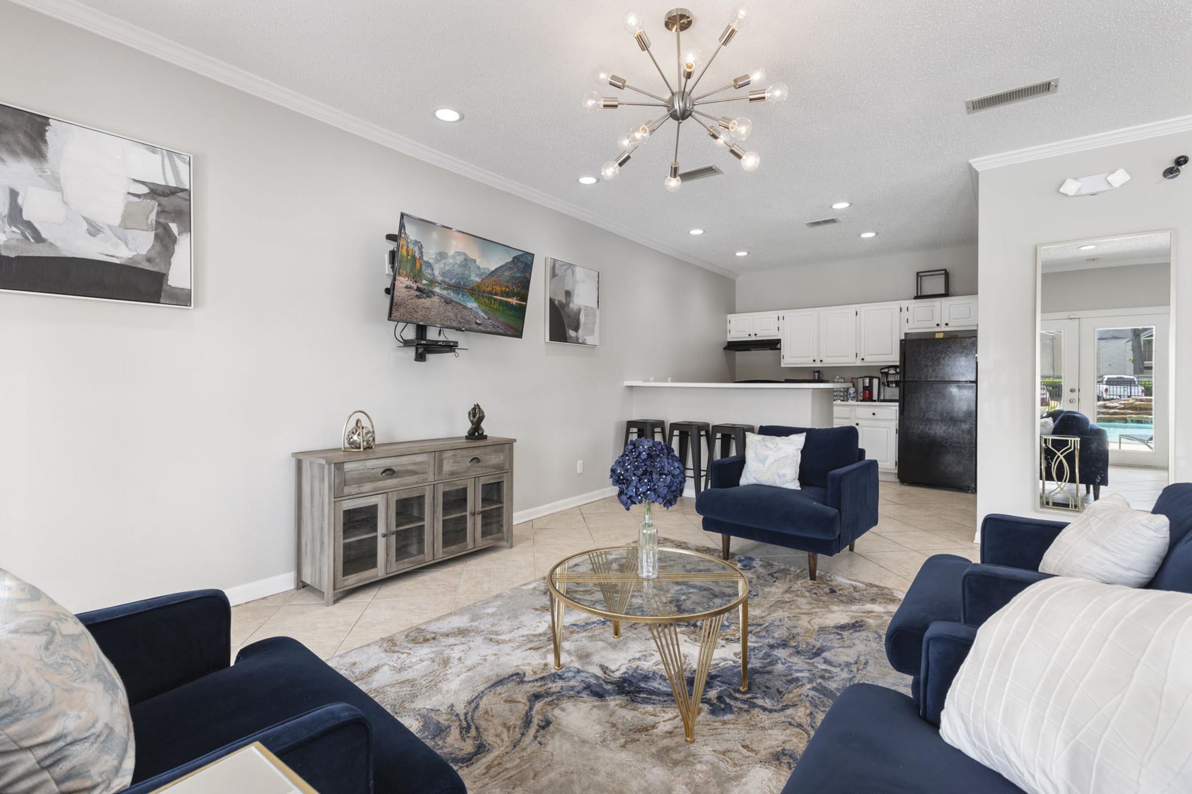 Living room interior featuring two blue accent chairs, a glass coffee table with a floral centerpiece, a mounted TV, and a dining area in the background. The space is well-lit, with modern decor and a neutral color palette, creating a welcoming atmosphere.