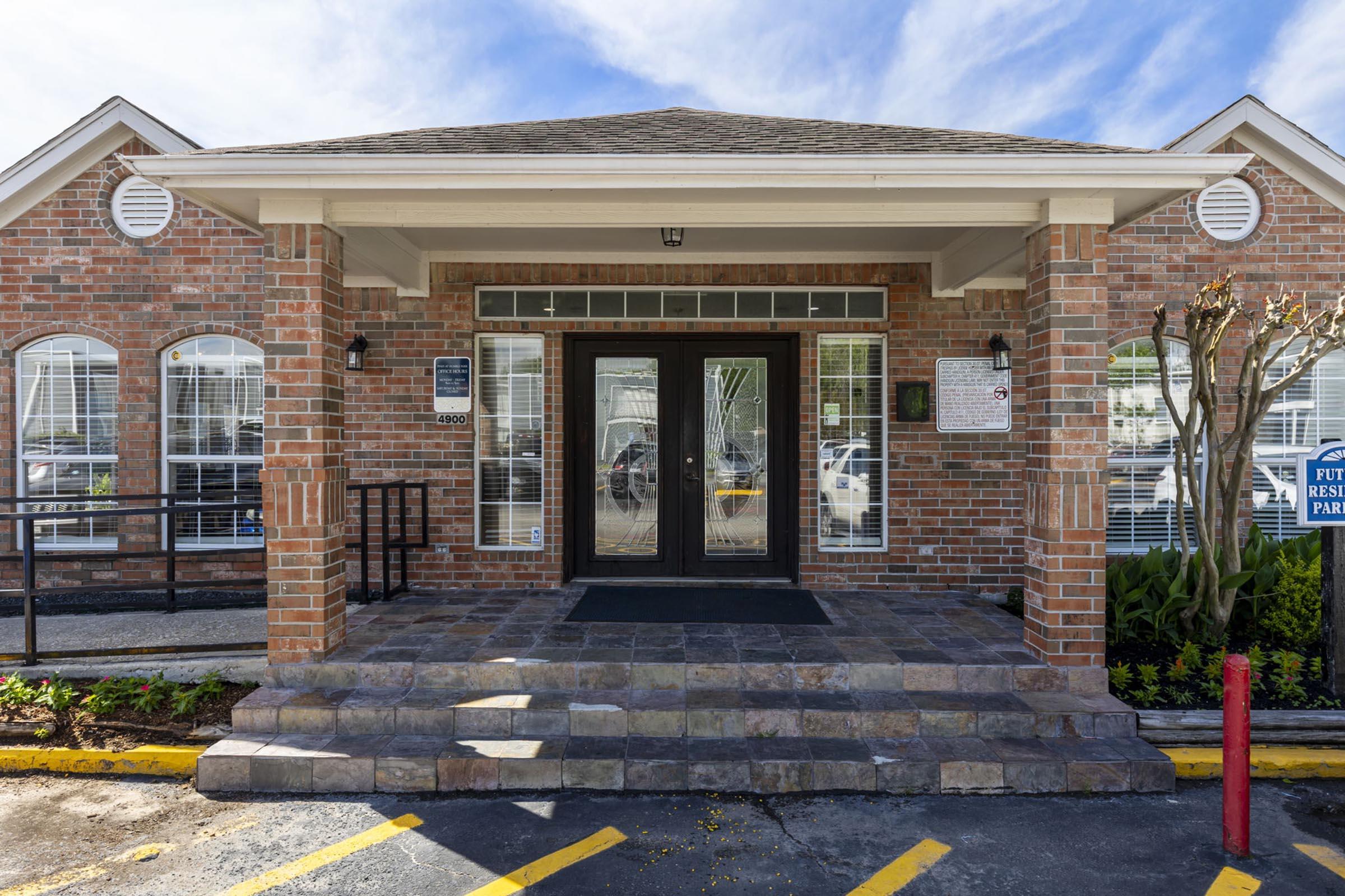 A brick building entrance featuring double glass doors, a flat roof with overhang, and large windows. The walkway is made of stone, leading to the doors. There are signs on the walls and a parking lot visible in the background. Green plants and a red parking barrier are in the foreground.