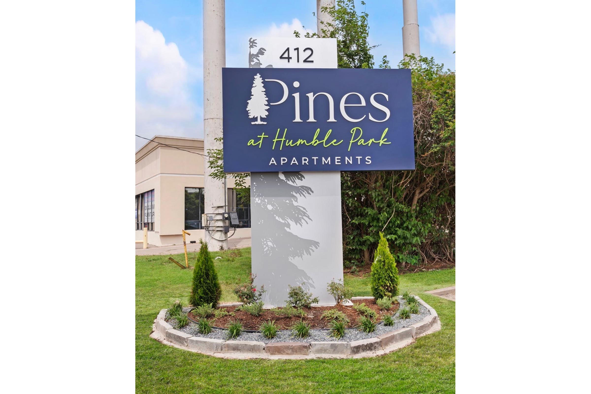 Sign for "Pines at Humble Park Apartments" featuring the number "412." The sign is prominently displayed with decorative landscaping including small shrubs and trees. The background shows a clear sky and an apartment building, suggesting a welcoming residential environment.