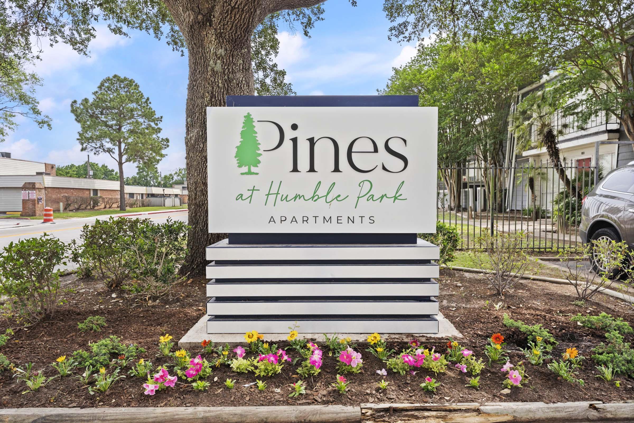 Sign for "Pines at Humble Park Apartments" featuring a modern design with green text and a tree graphic, surrounded by colorful flowers and a landscaped area. In the background, there are trees and a glimpse of nearby buildings under a clear blue sky.