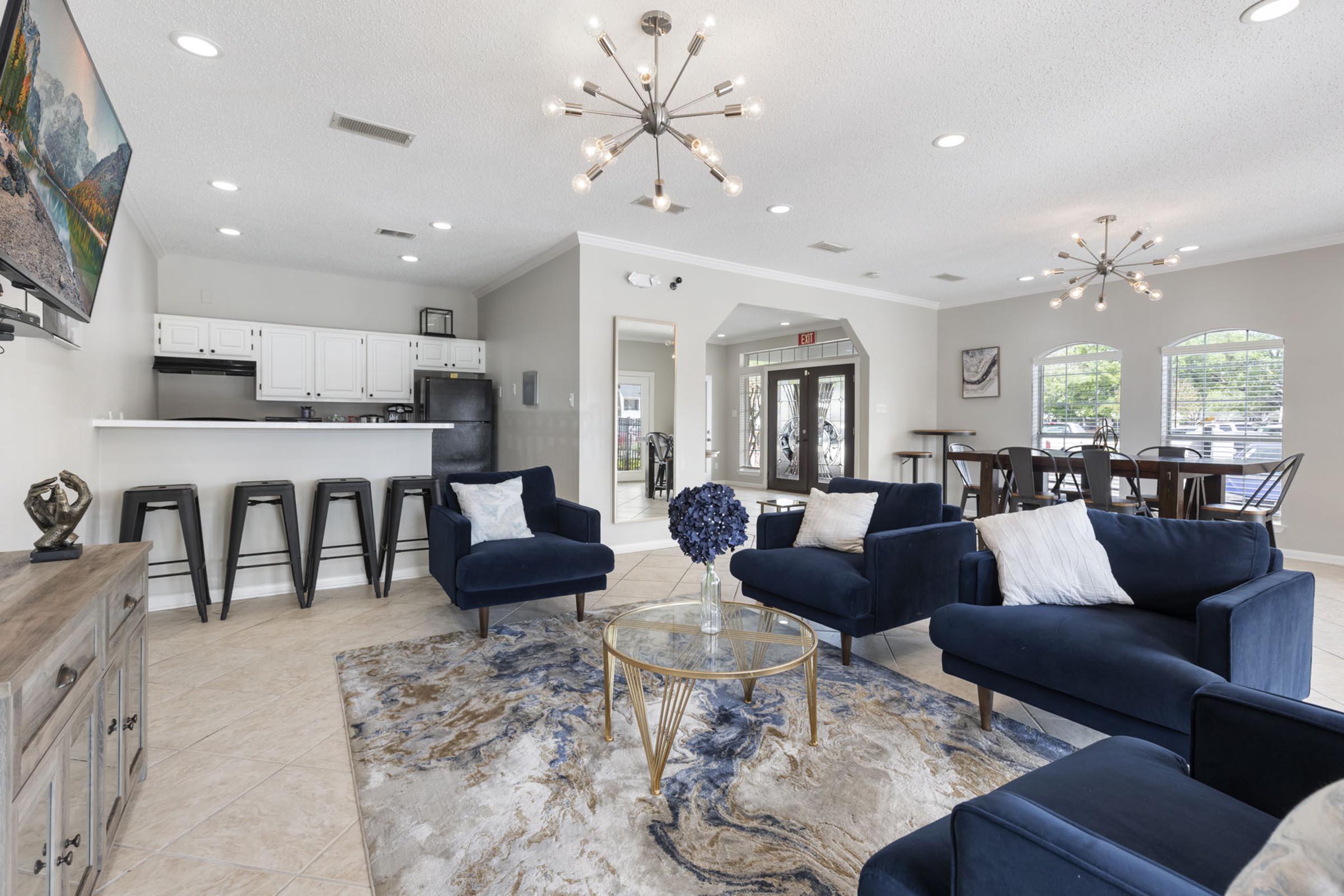 A spacious and modern living area featuring navy blue sofas, a glass coffee table, and a decorative rug. In the background, a kitchen area with white cabinets and bar stools is visible. Large windows allow natural light to brighten the space. Overall, the room is well-decorated and inviting.