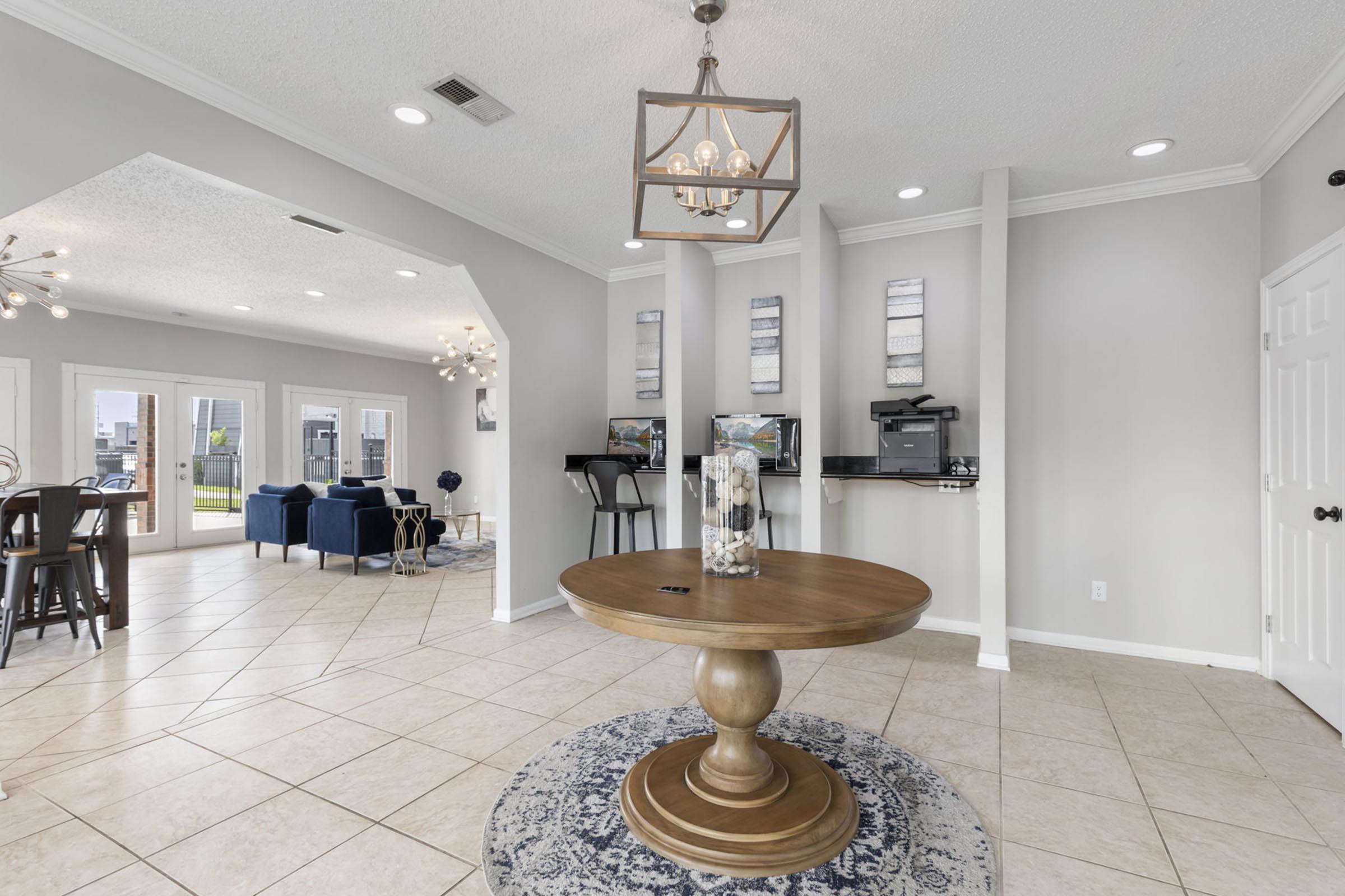 Interior view of a modern, spacious room with a round wooden table at the center, a decorative vase on top, and a patterned rug beneath. The room features neutral-colored walls, tiled flooring, and bright lighting. In the background, there are dark blue chairs, a small kitchen area, and large windows allowing natural light.