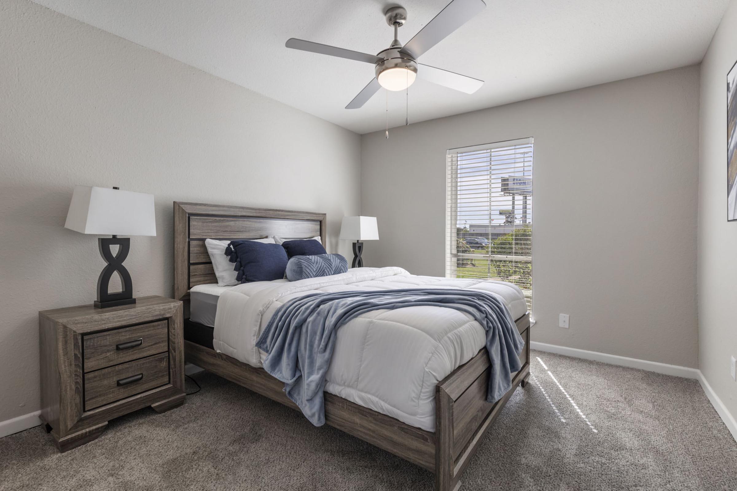 A cozy bedroom featuring a wooden bed with white bedding and blue pillows. There are two bedside lamps on wooden nightstands, and a ceiling fan overhead. A window lets in natural light, showing a view outside. The walls are painted a light color, and the carpet is soft and neutral.