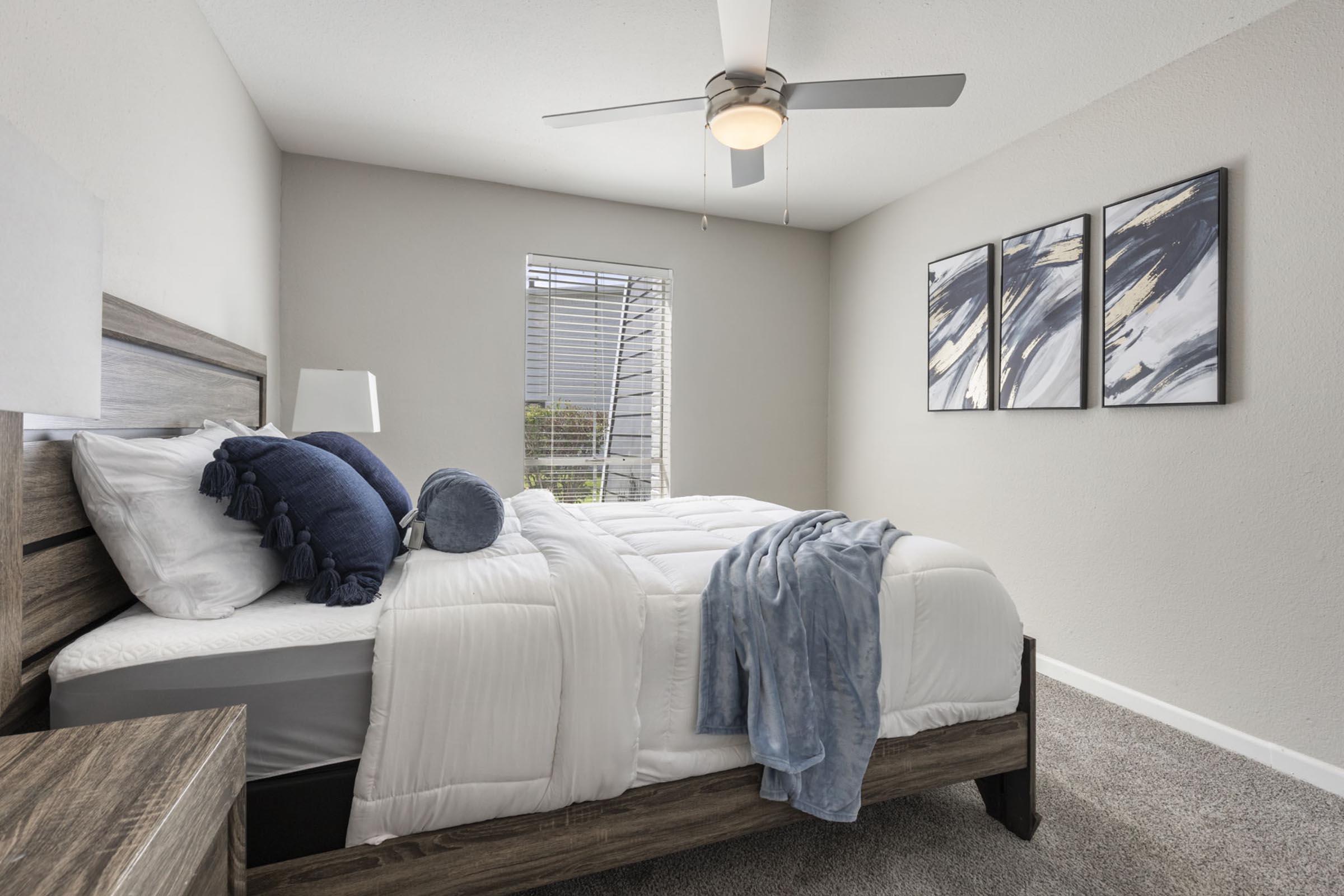 A modern bedroom featuring a neatly made bed with white linens, a blue throw blanket, and accent pillows. There is a nightstand with a lamp, a window with shades, and three abstract art pieces on the wall. A ceiling fan is visible, adding a contemporary touch to the cozy space.