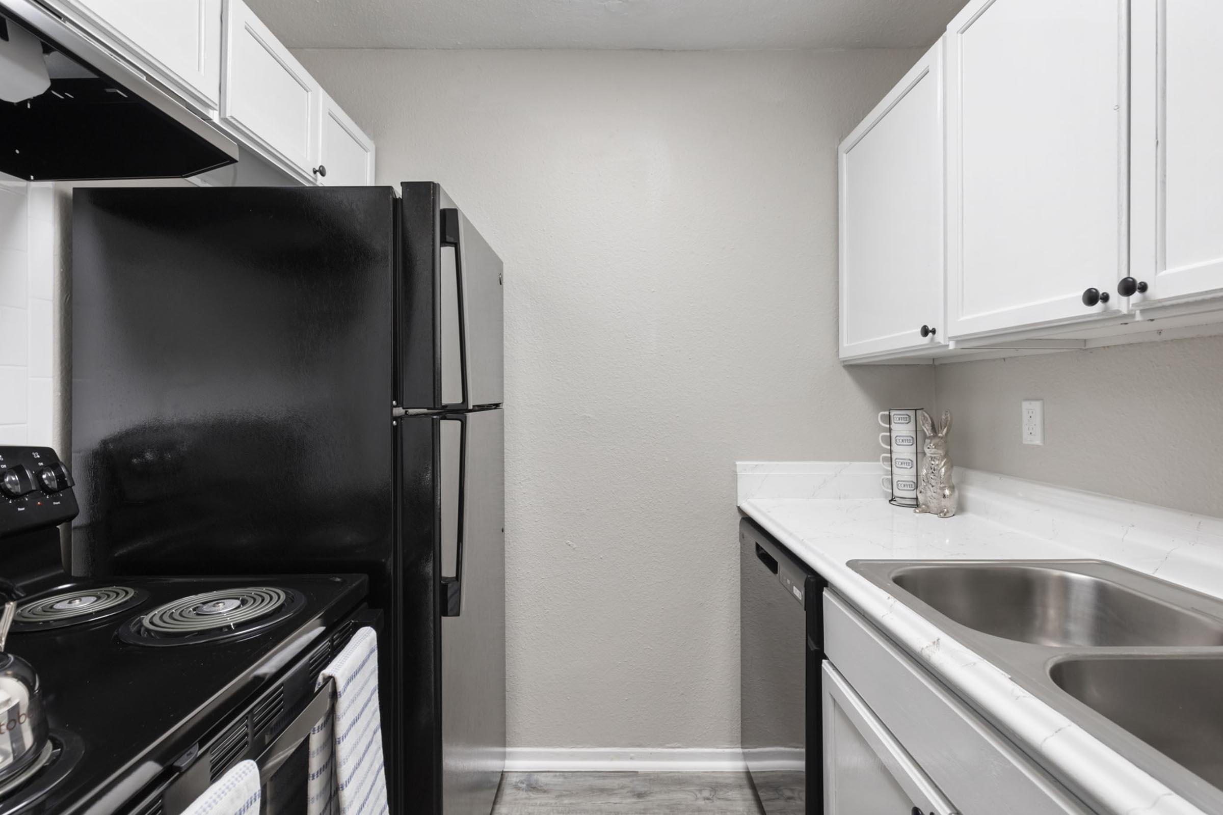 A modern kitchen featuring white cabinets, black appliances including a stove and refrigerator, and a double sink. The walls are light-colored, and the countertop is white with a subtle texture. The space is well-organized and clean, with a few decorative items on the countertop.