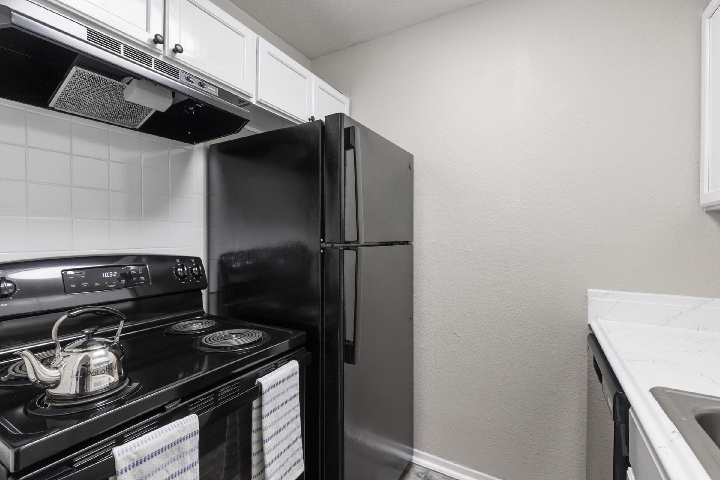 A small kitchen featuring a black fridge and stove, white cabinetry, and gray walls. The stove has a kettle on it, and there are dish towels hanging from the oven. A sink is visible on the right, with a countertop providing additional workspace. The overall decor is modern and minimalistic.