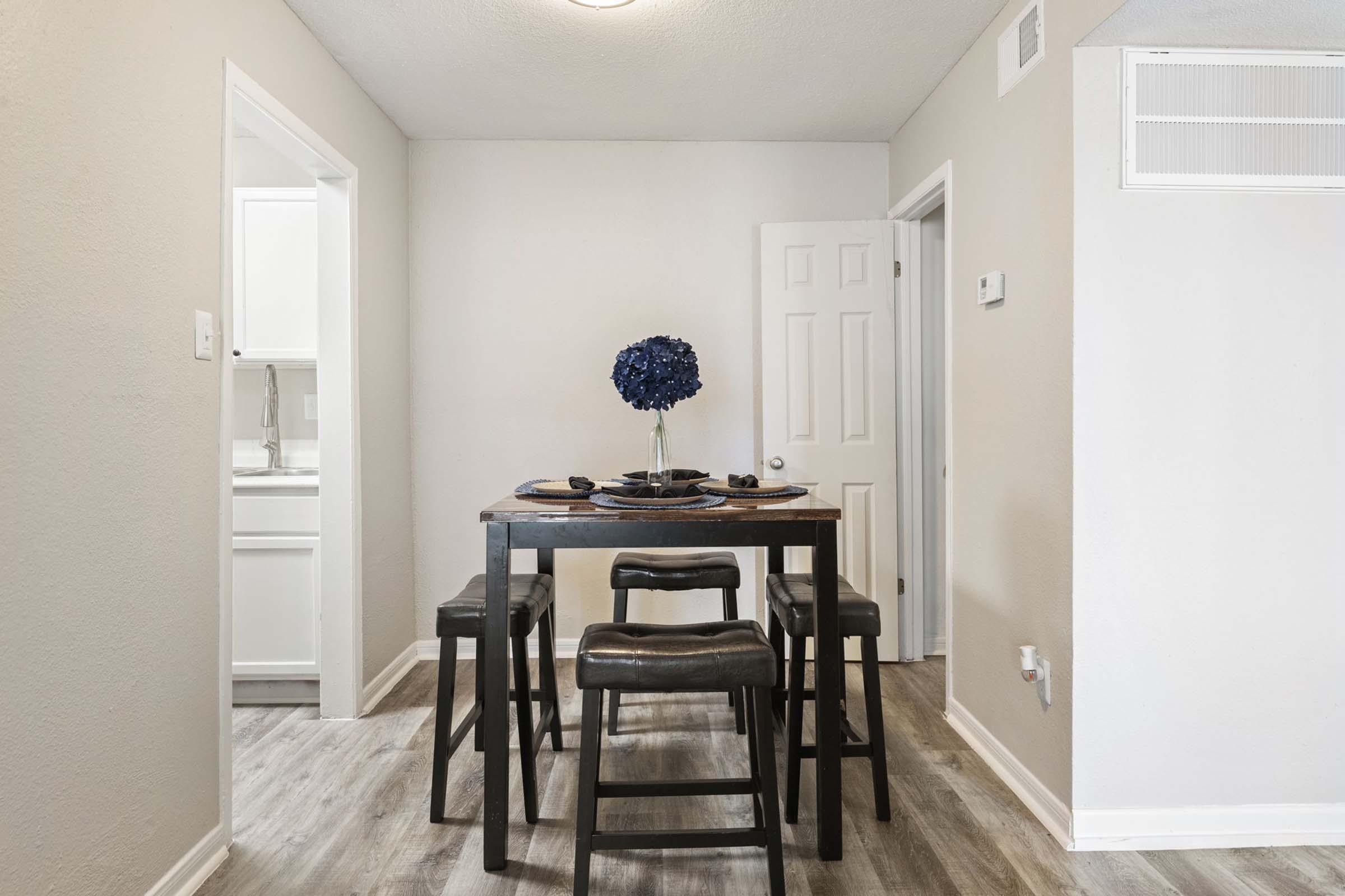 A modern dining area featuring a wooden table with dark seating and a blue floral centerpiece. The walls are painted in a light shade, and there are doorways leading to other rooms, with a light-colored floor adding to the contemporary feel of the space.