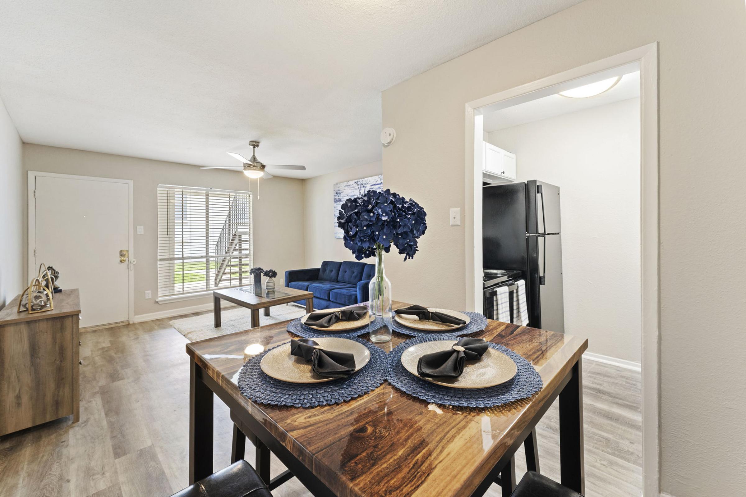 Modern living space featuring a dining table set for two with a blue floral centerpiece. In the background, there's a cozy blue sofa and a doorway leading to a kitchen area with a black refrigerator. Natural light filters in through vertical blinds. The décor is neutral and inviting.