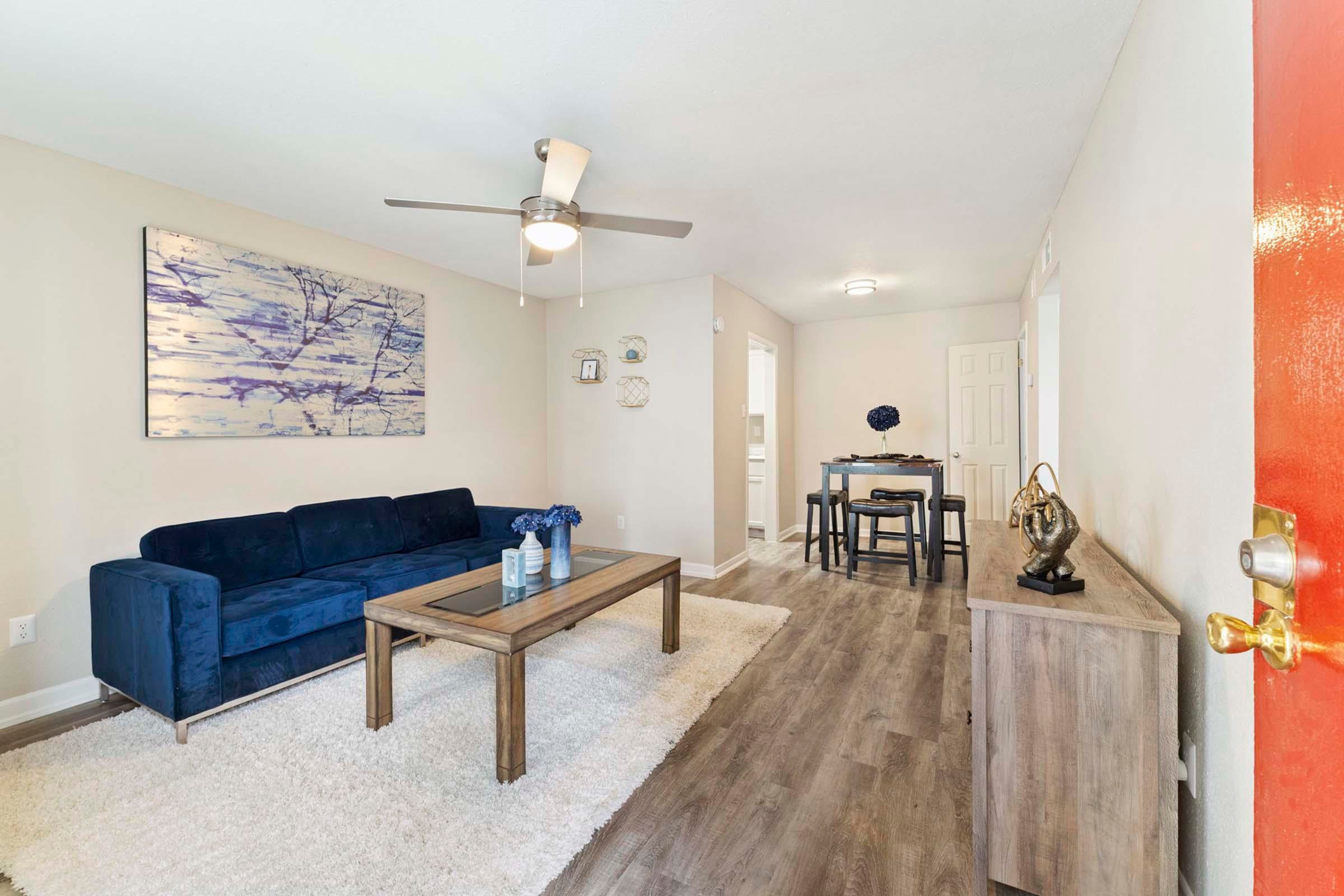 A modern living room featuring a blue sofa, a wooden coffee table, and decorative items. The walls are a neutral color, and light-colored flooring complements the space. A dining area with a small table is visible in the background. A ceiling fan is present, and a colorful piece of artwork adorns the wall.