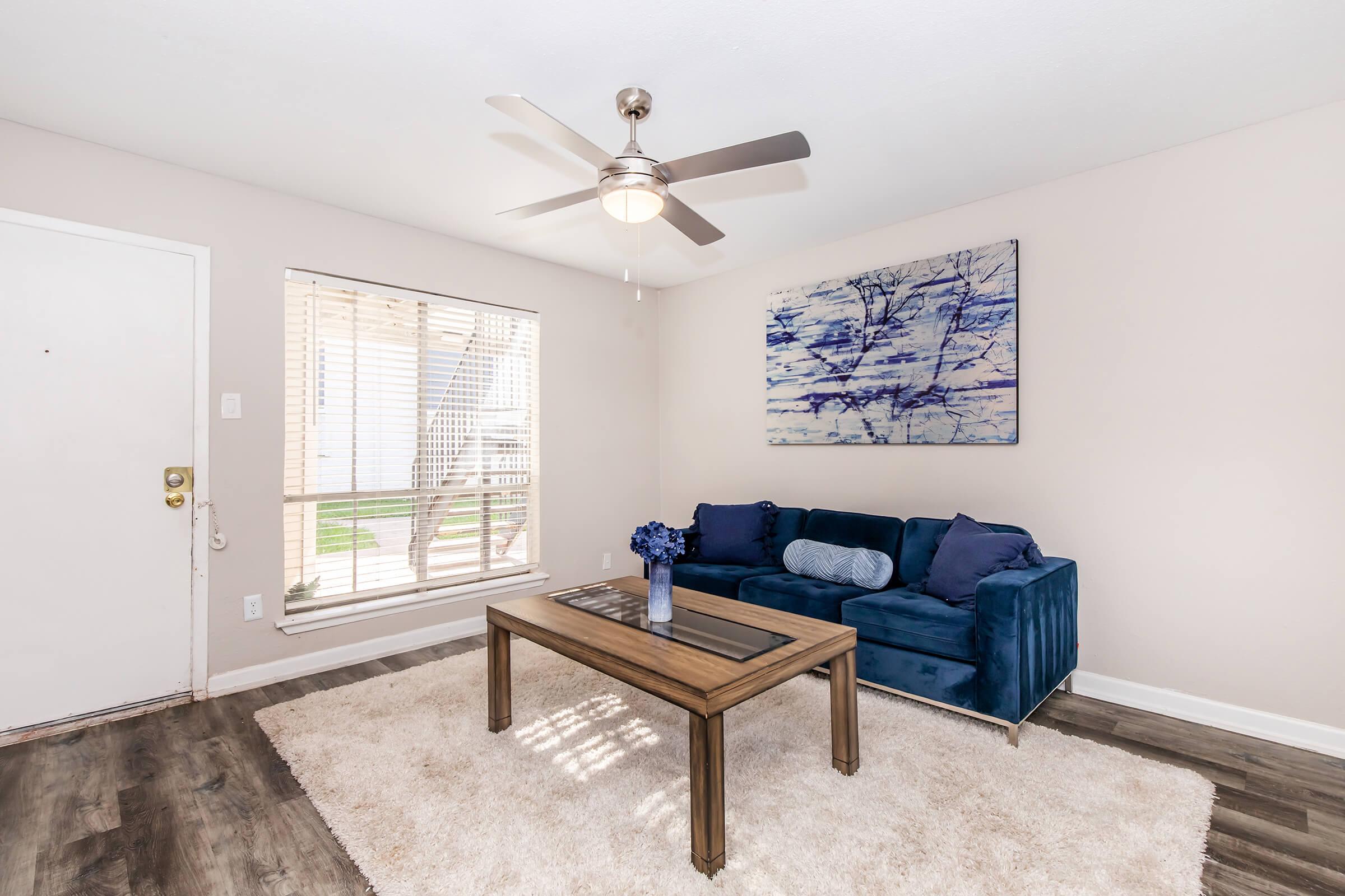 A cozy living room featuring a navy blue sofa with blue pillows, a wooden coffee table, and a light-colored rug. There is a ceiling fan and natural light coming through a window with blinds. A modern artwork is displayed on the wall above the sofa, adding a decorative touch to the space.