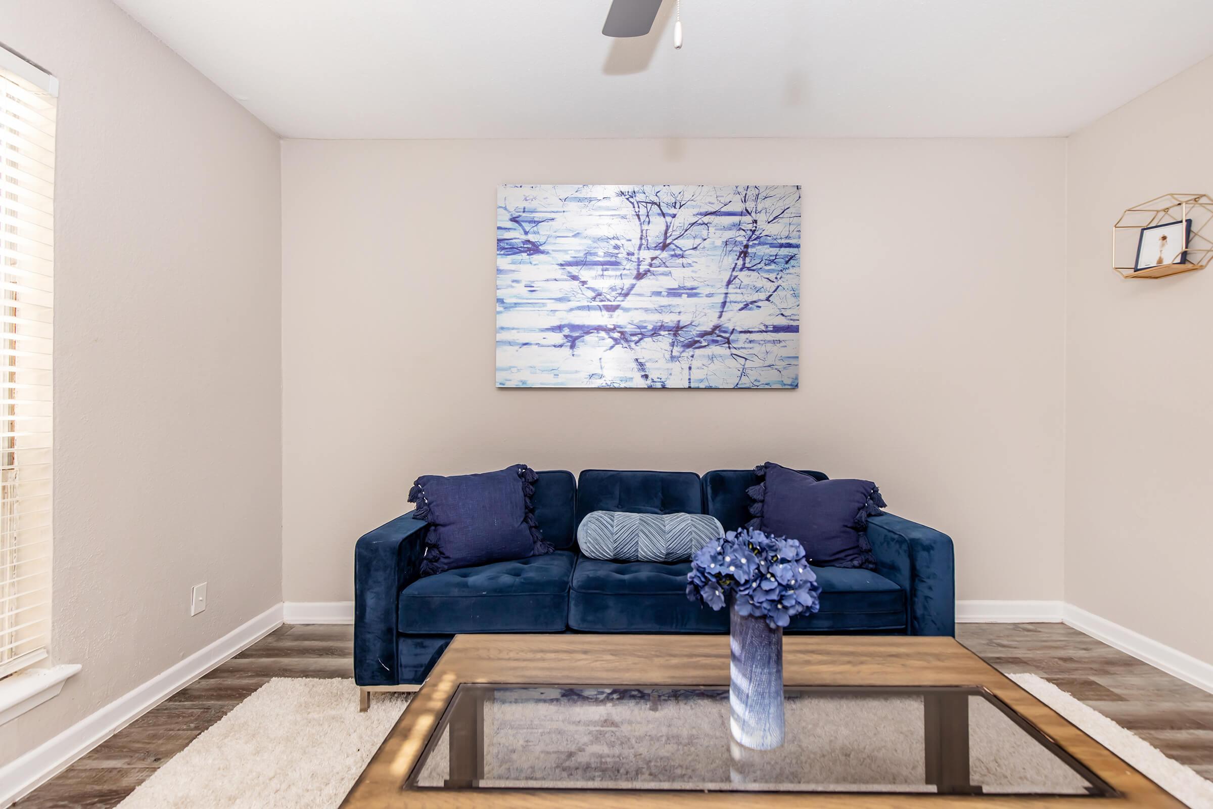 A cozy living room featuring a deep blue sofa with decorative pillows, a wooden coffee table with a glass top, and a vase of purple flowers. On the wall, there is a light blue abstract painting. Natural light streams in through a window with blinds, creating a warm atmosphere.