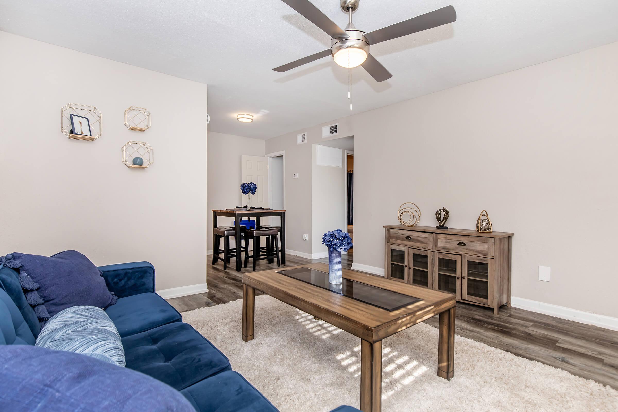 A cozy living room featuring a blue sofa with decorative pillows, a wooden coffee table, and a light-colored area rug. There is a small dining table in the background, a ceiling fan overhead, and decorative items on a console table. The walls are painted in a neutral tone, creating a welcoming atmosphere.
