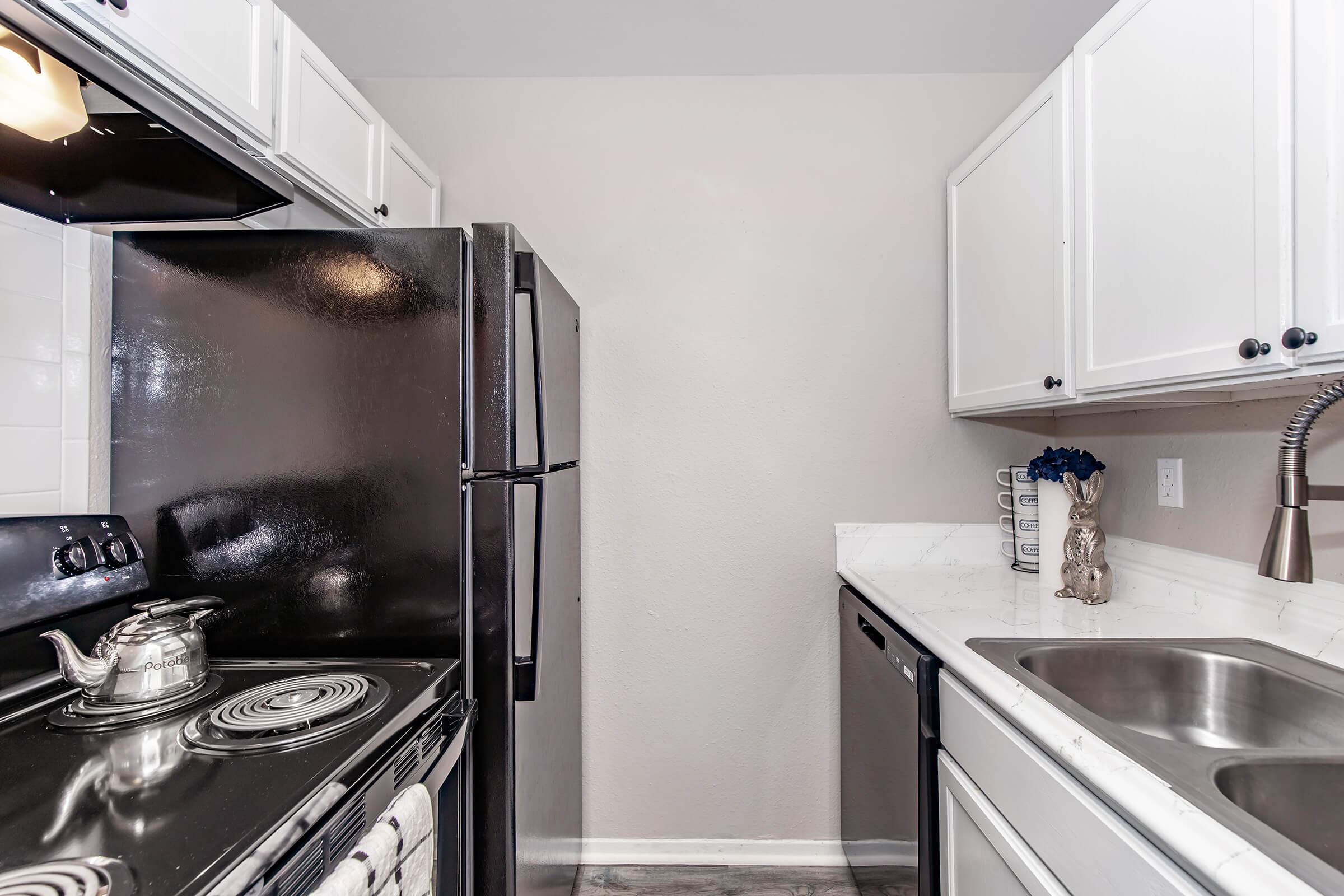 A modern kitchen featuring white cabinets, black appliances including a fridge and stove, a stainless steel sink, and a marble countertop. The room has gray walls and is neatly organized, with a kettle on the stove and decorative items on the counter.