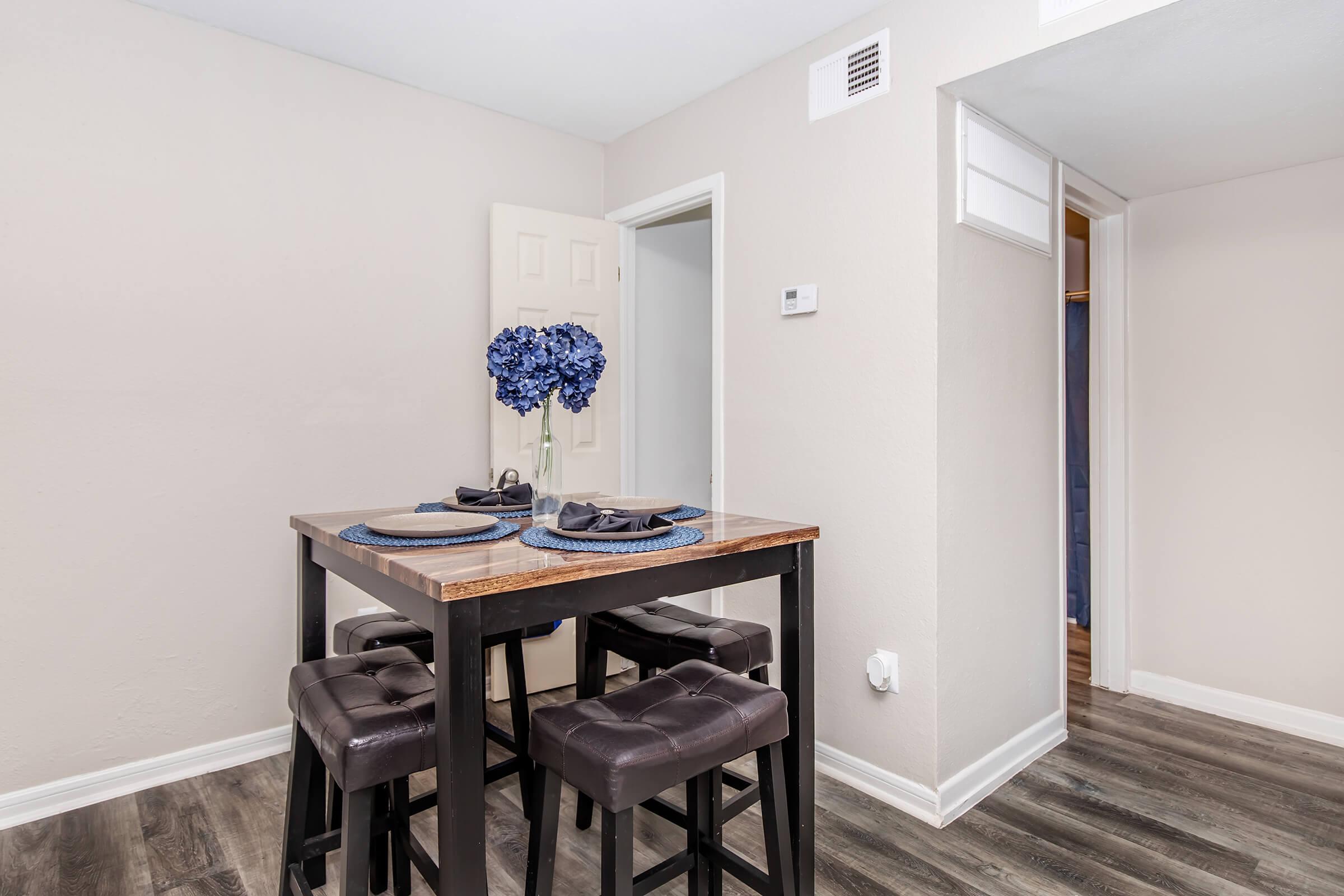 A small dining area featuring a wooden table with blue placemats and two dark upholstered stools. A vase with blue flowers sits in the center of the table. Light-colored walls and a doorway in the background create a bright, modern atmosphere.