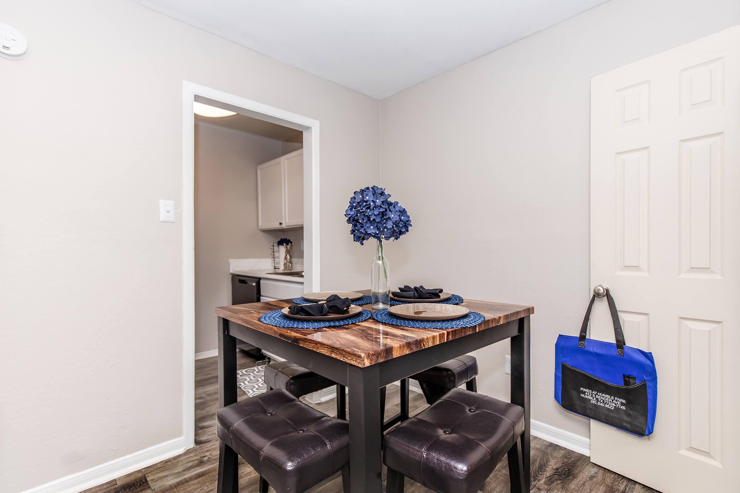 A cozy dining area featuring a wooden table set with blue placemats, black plates, and a bouquet of blue flowers in a vase. Two dark stools are at the table, and a closed door leads to another room in the background. A blue bag hangs on the door. The walls are light gray, creating a warm atmosphere.