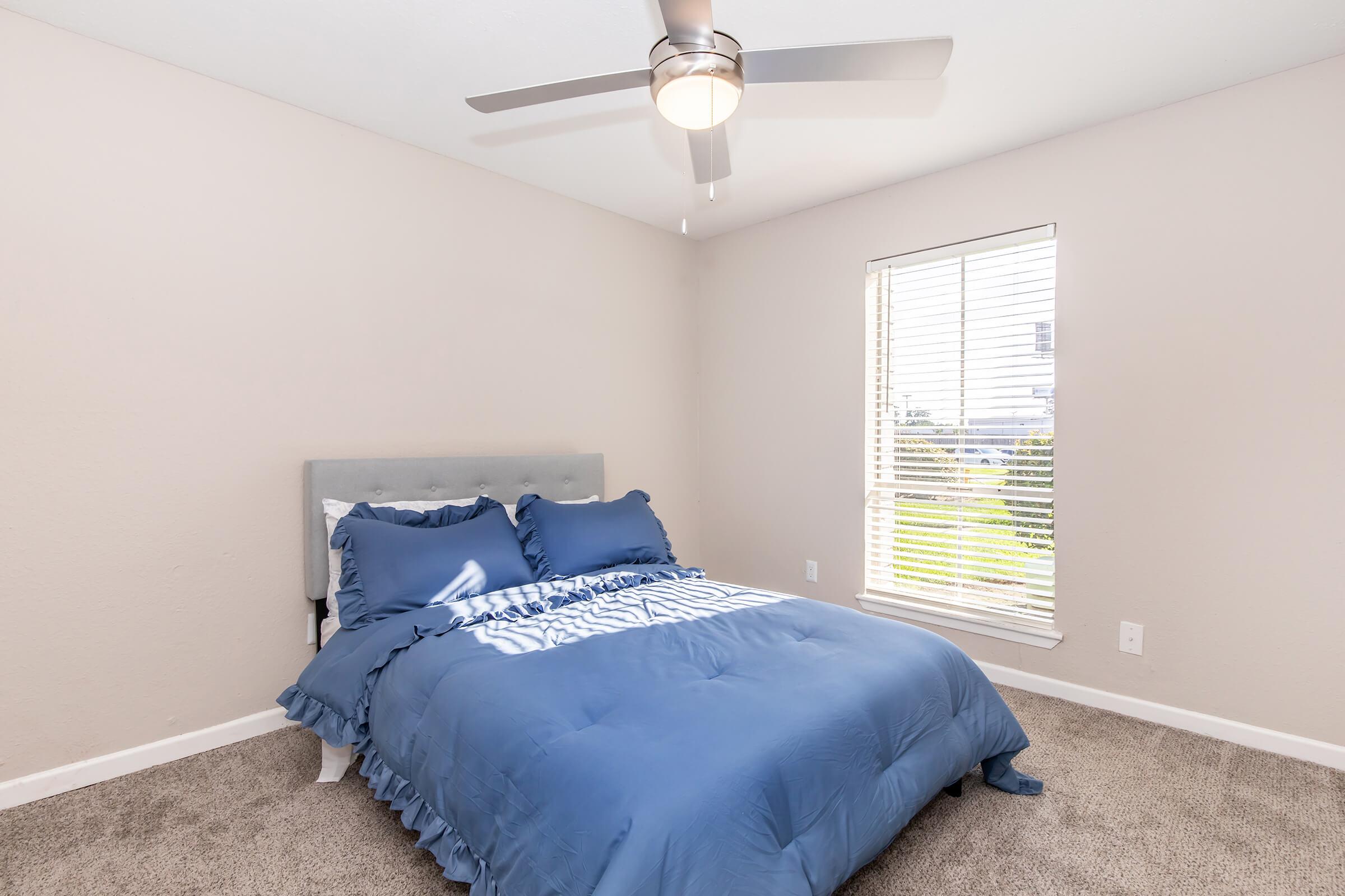 A well-lit bedroom featuring a queen-sized bed with a blue ruffled comforter and matching pillows. The room has light beige walls and a large window with white blinds, allowing natural light to enter. A ceiling fan is installed above, and the carpeted floor adds warmth to the space.