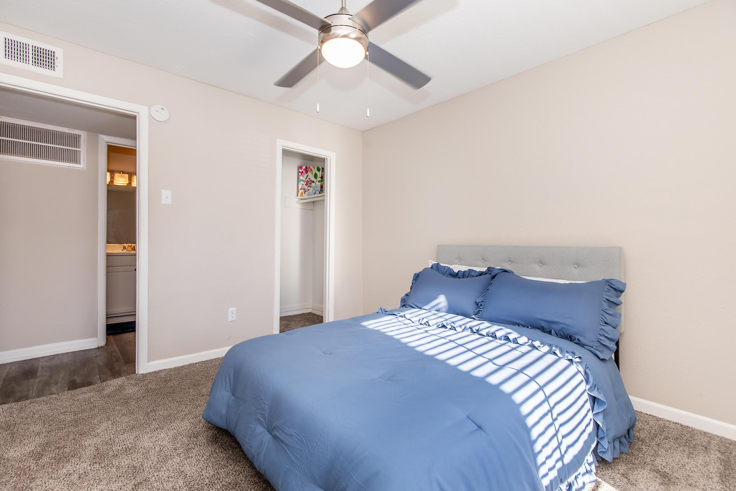 A cozy bedroom featuring a queen-sized bed with blue bedding and a grey upholstered headboard. Sunlight streams through a window, casting striped shadows on the carpet. A ceiling fan is above, and there is a doorway leading to a bathroom. A small closet is visible on the side. The walls are painted a soft beige.