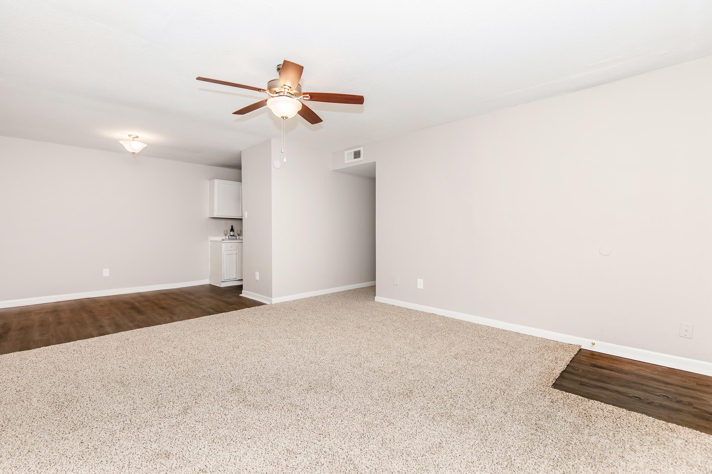 A spacious, well-lit living area with light beige carpet and a ceiling fan. The walls are painted in a soft neutral color, and a doorway leads to a small kitchenette in the background. The room is empty, creating an open and inviting atmosphere.