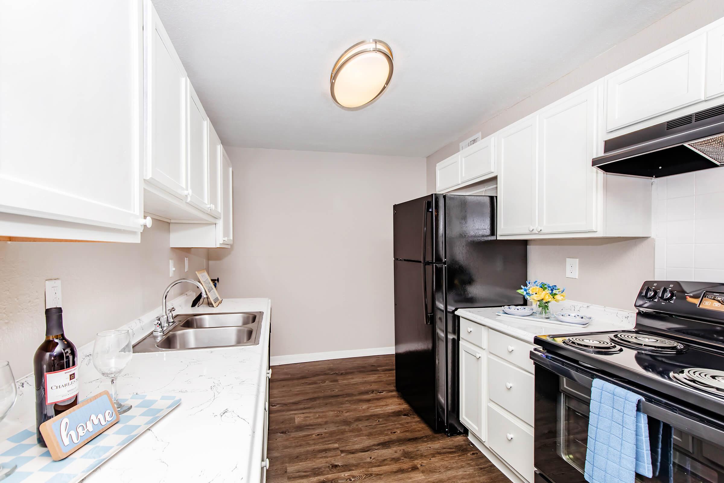 Modern kitchen featuring white cabinets, a black refrigerator, and stainless steel appliances. The countertop has a marble design, and there's a bottle of wine with a glass beside it. A blue dish towel is hanging on the stove, and a vase with yellow flowers adds a pop of color. Bright overhead lighting enhances the space.