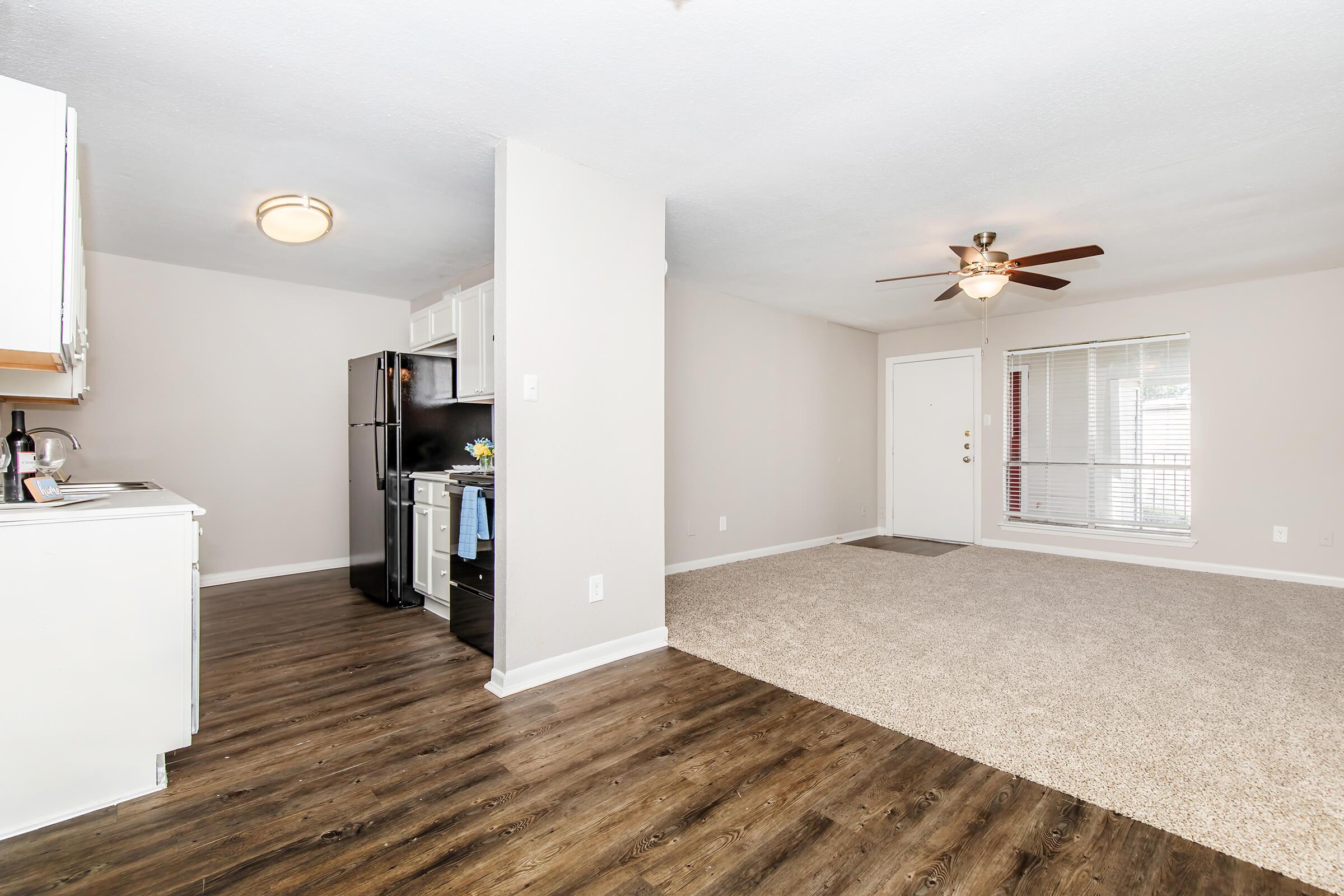 A spacious living area featuring a modern kitchen with white cabinets and stainless steel appliances. The room has light-colored walls, a ceiling fan, and a doorway leading to an entrance. Natural light comes from a window with blinds, and the floor is a combination of laminate and carpet.
