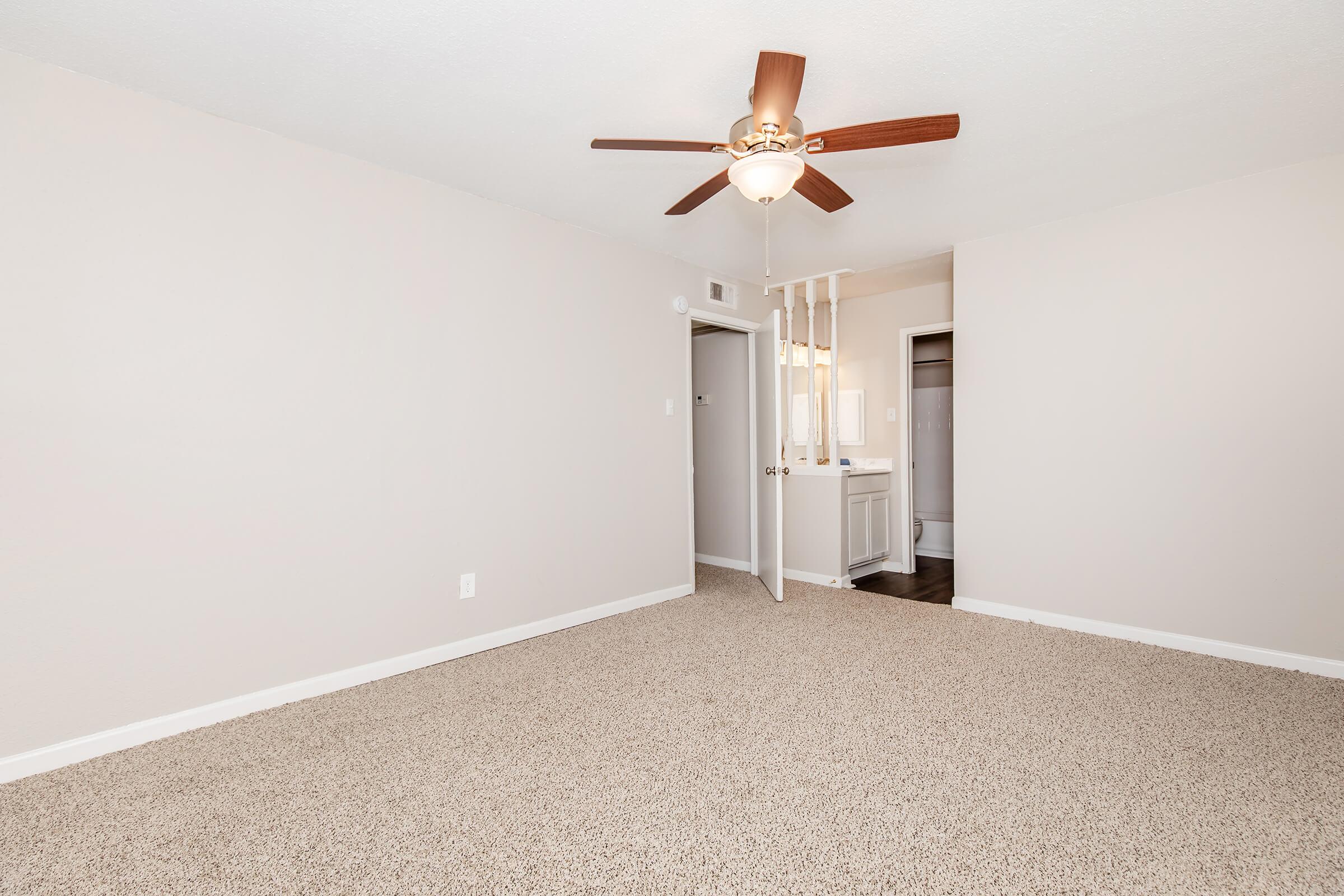 A spacious room featuring a ceiling fan with wooden blades, light beige walls, and plush carpeting. An open doorway leads to a bathroom area, and there’s a closet visible in the background. The room is well-lit, creating a cozy atmosphere.