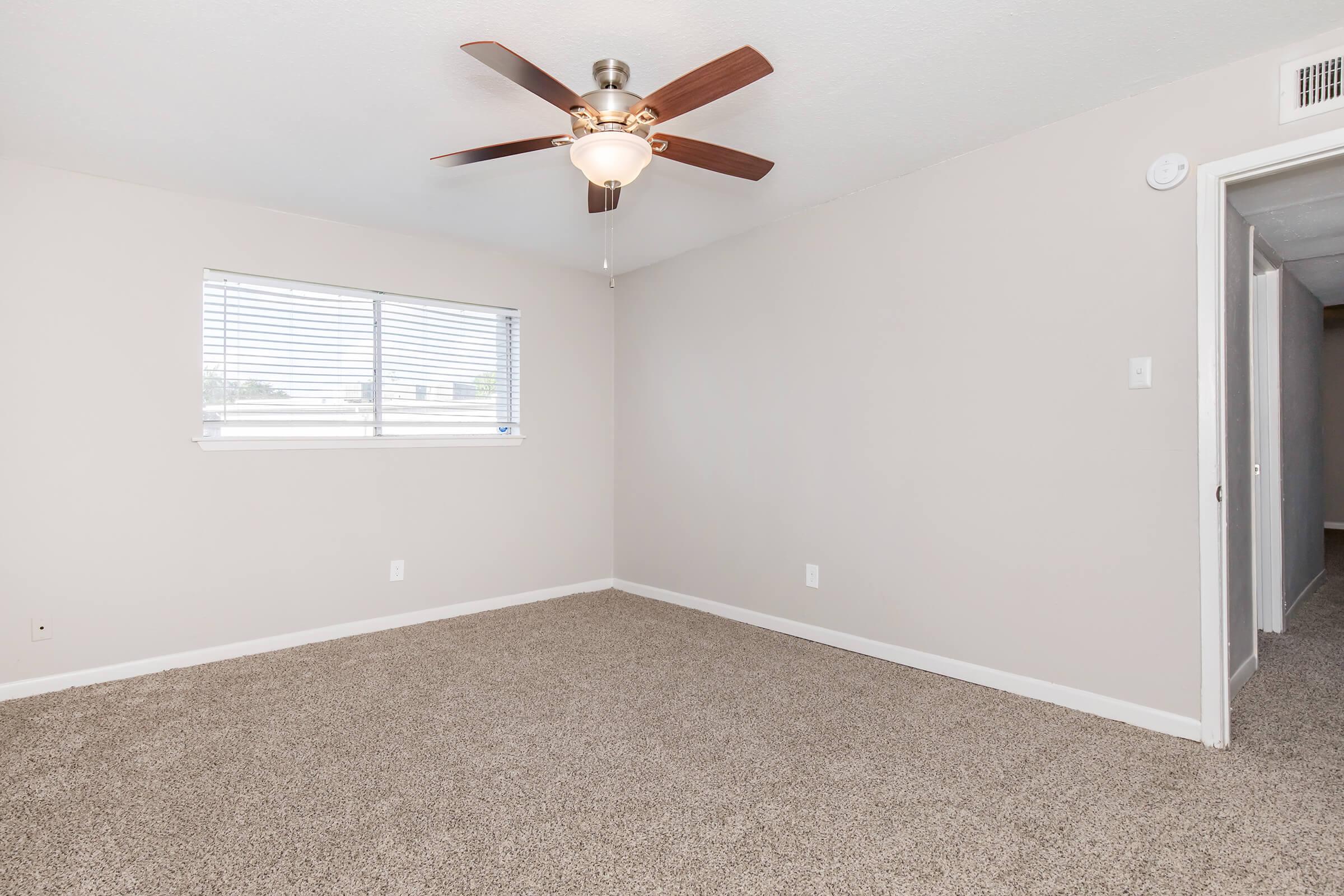 A spacious room with light beige walls and carpeted flooring. It features a ceiling fan with a light fixture, a window with blinds allowing natural light, and a door leading to a hallway. The room is unfurnished, creating an open and airy atmosphere.