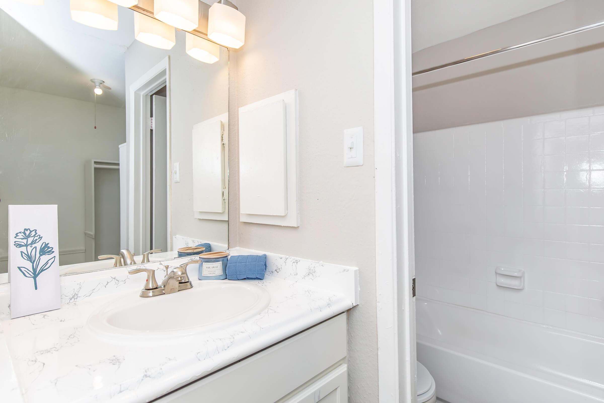 A clean, bright bathroom featuring a white countertop with a sink, a modern mirror, and a light fixture above. There is a decorative flower illustration on the counter, a small blue towel, and a bathtub with a shower rod in the background. The walls are painted in a light color, enhancing the space's openness.