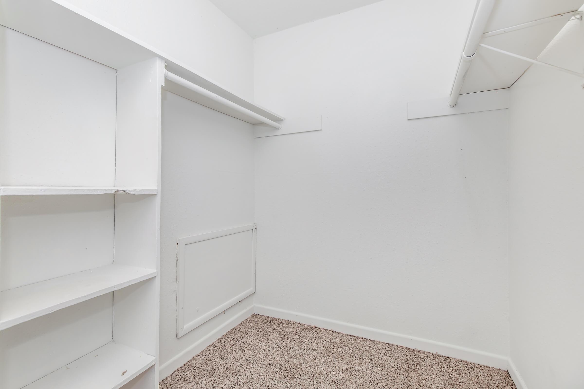 A small, empty room with white walls and carpeted flooring. There are built-in shelves on one side and a hanging rod for clothes on the opposite wall. The space appears clean and minimally furnished, suitable for storage or as a closet.