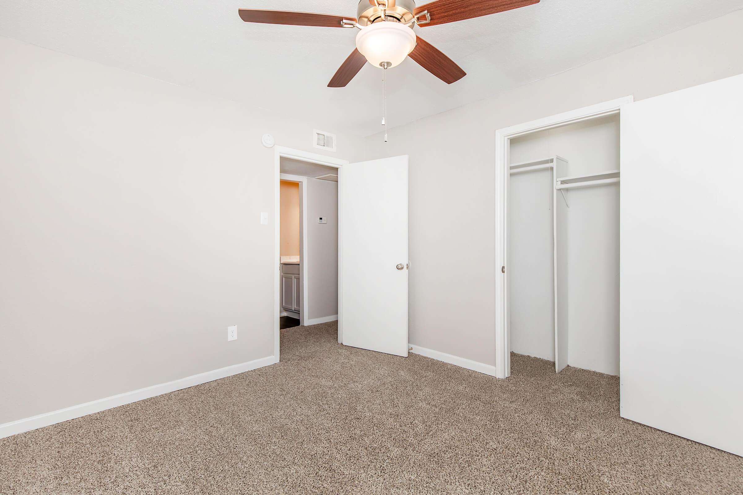 A well-lit, neutral-colored bedroom featuring a ceiling fan, freshly painted walls, and carpeted flooring. The room includes an open door leading to a closet on one side and another door opening to an adjoining area, possibly a bathroom or hallway. The space appears clean and inviting.