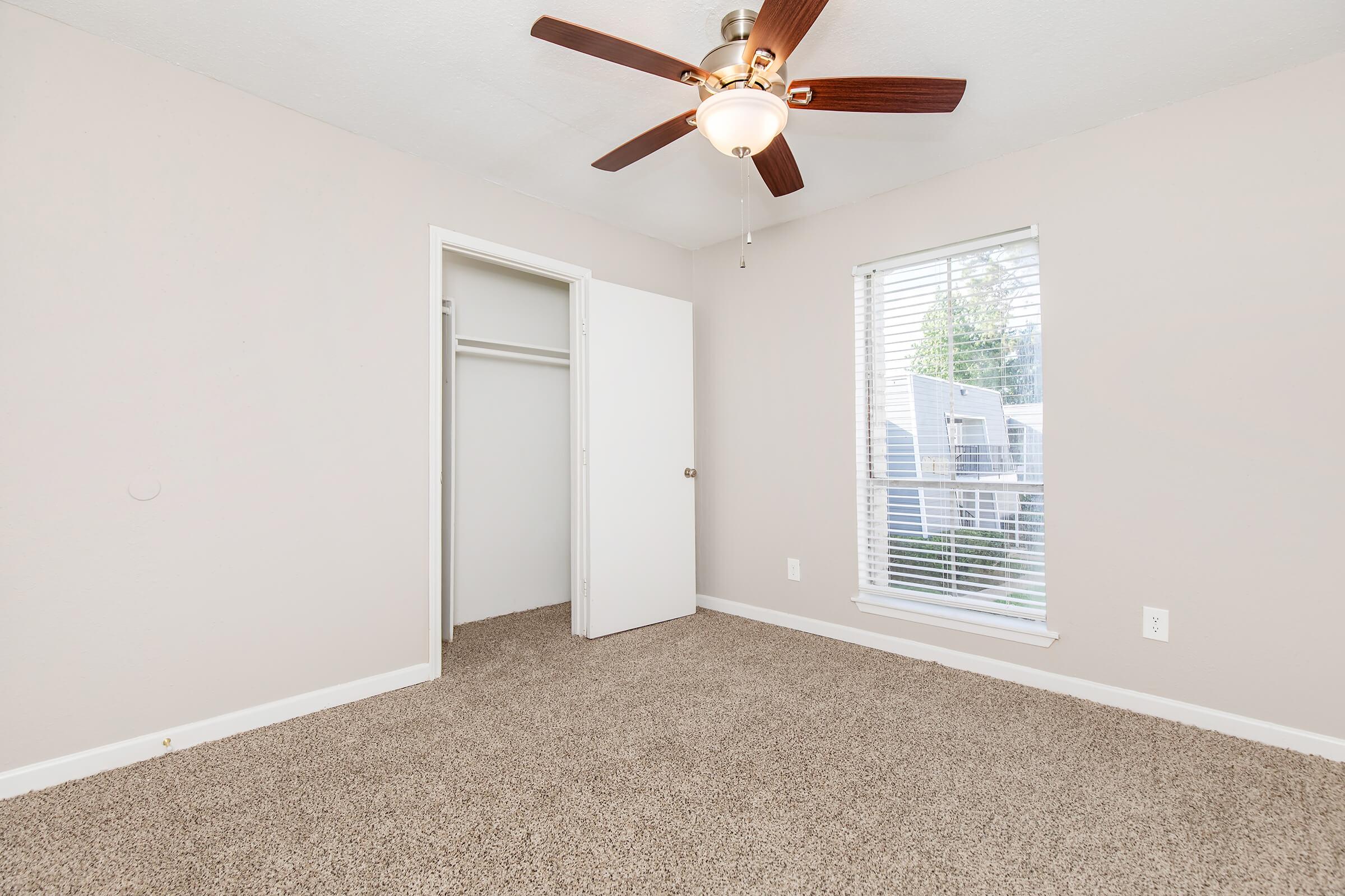 A well-lit, empty bedroom featuring light beige walls and a ceiling fan. There is a window with blinds allowing natural light in, and a spacious closet with white doors. The floor is covered in soft, brown carpet, creating a cozy atmosphere.