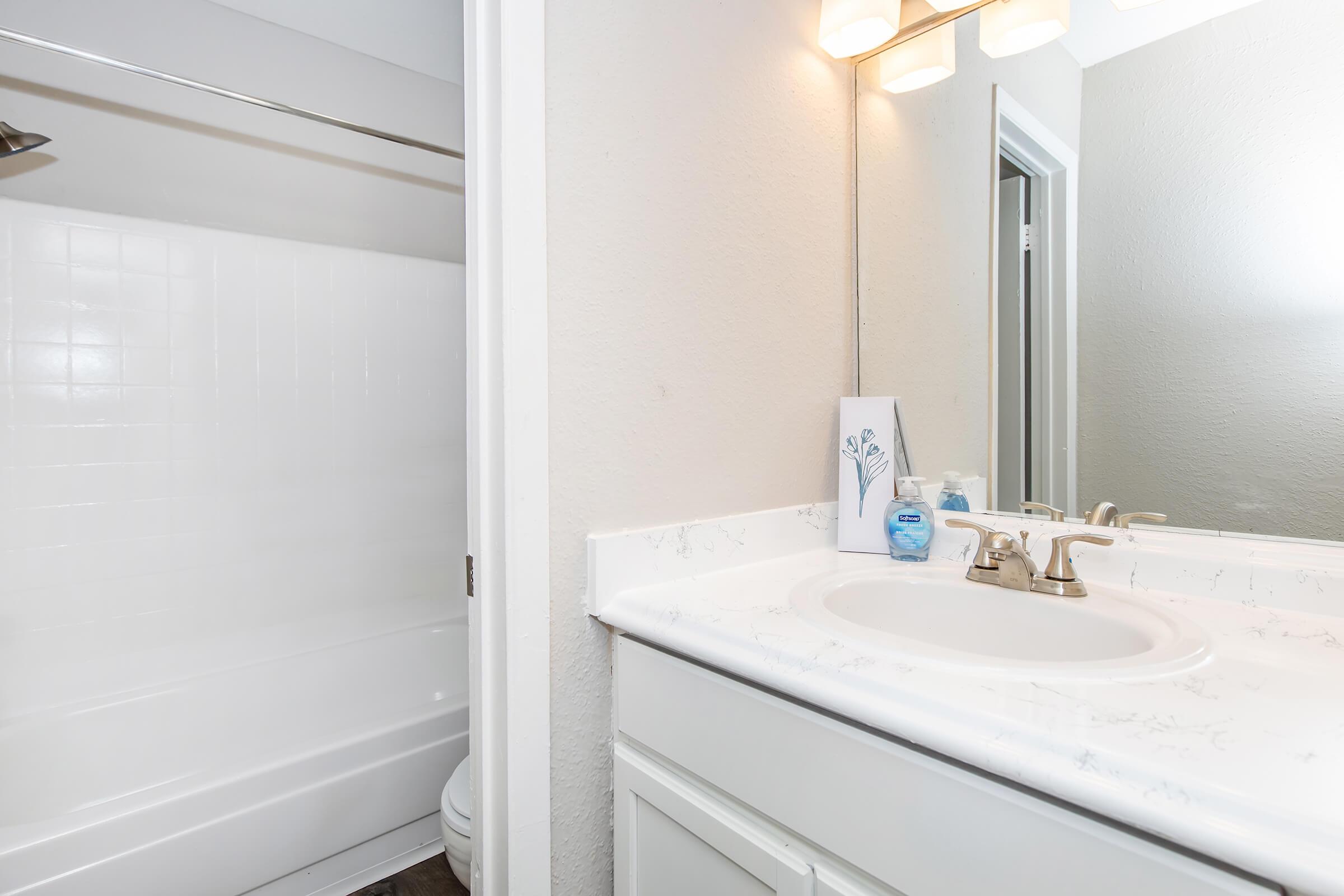 A clean bathroom featuring a white sink with faucet and soap dispenser, a mirror, and a shower area with a white tub. The walls are light-colored, and the floor is dark. The overall ambiance is neat and modern, ideal for a relaxing space.