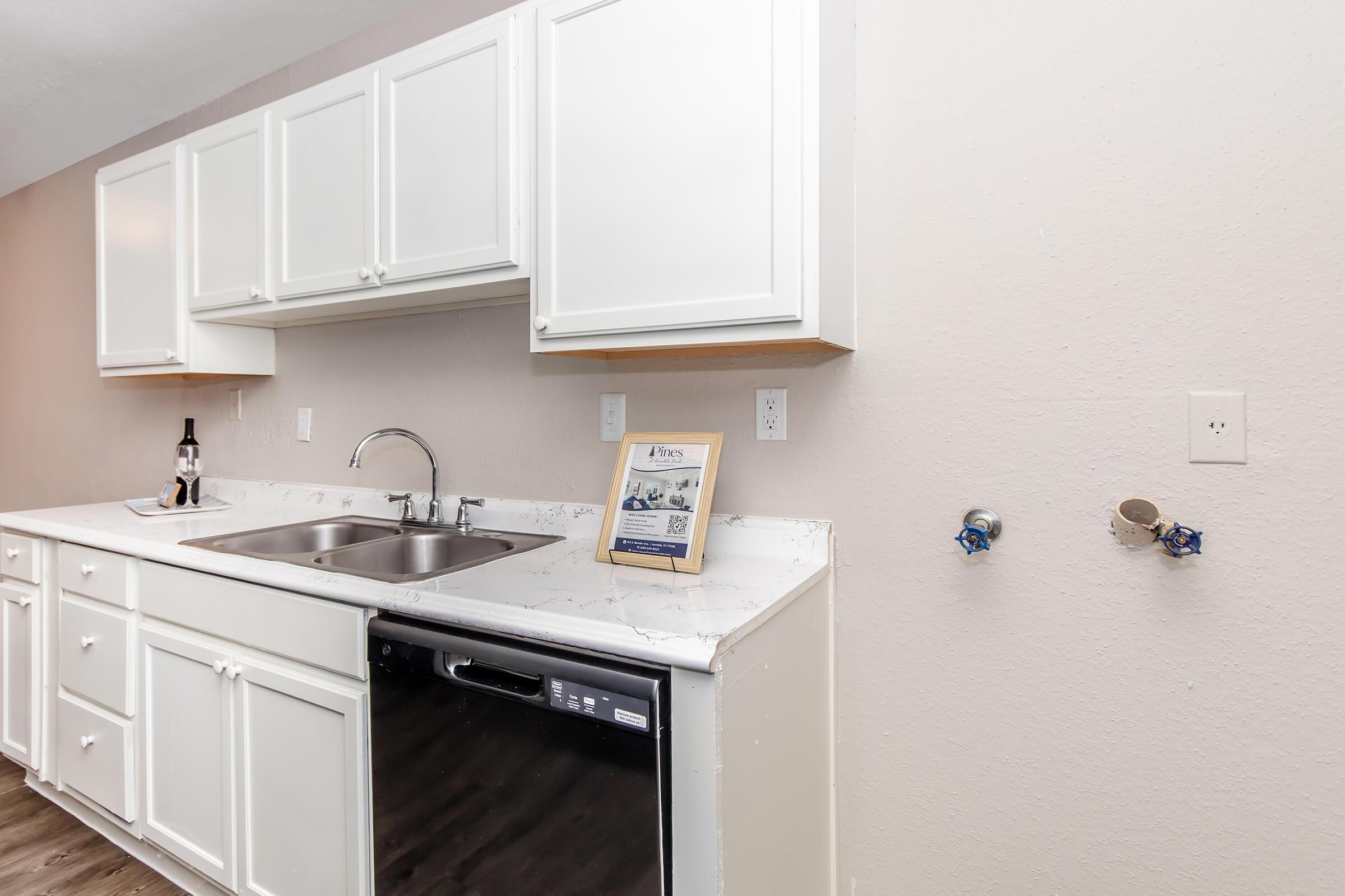 A modern kitchen featuring white cabinetry, a double sink with a sleek faucet, and a dishwasher situated underneath the counter. A decorative sign is placed on the countertop. The wall is painted a soft color, and there are two water connections visible on the right side.
